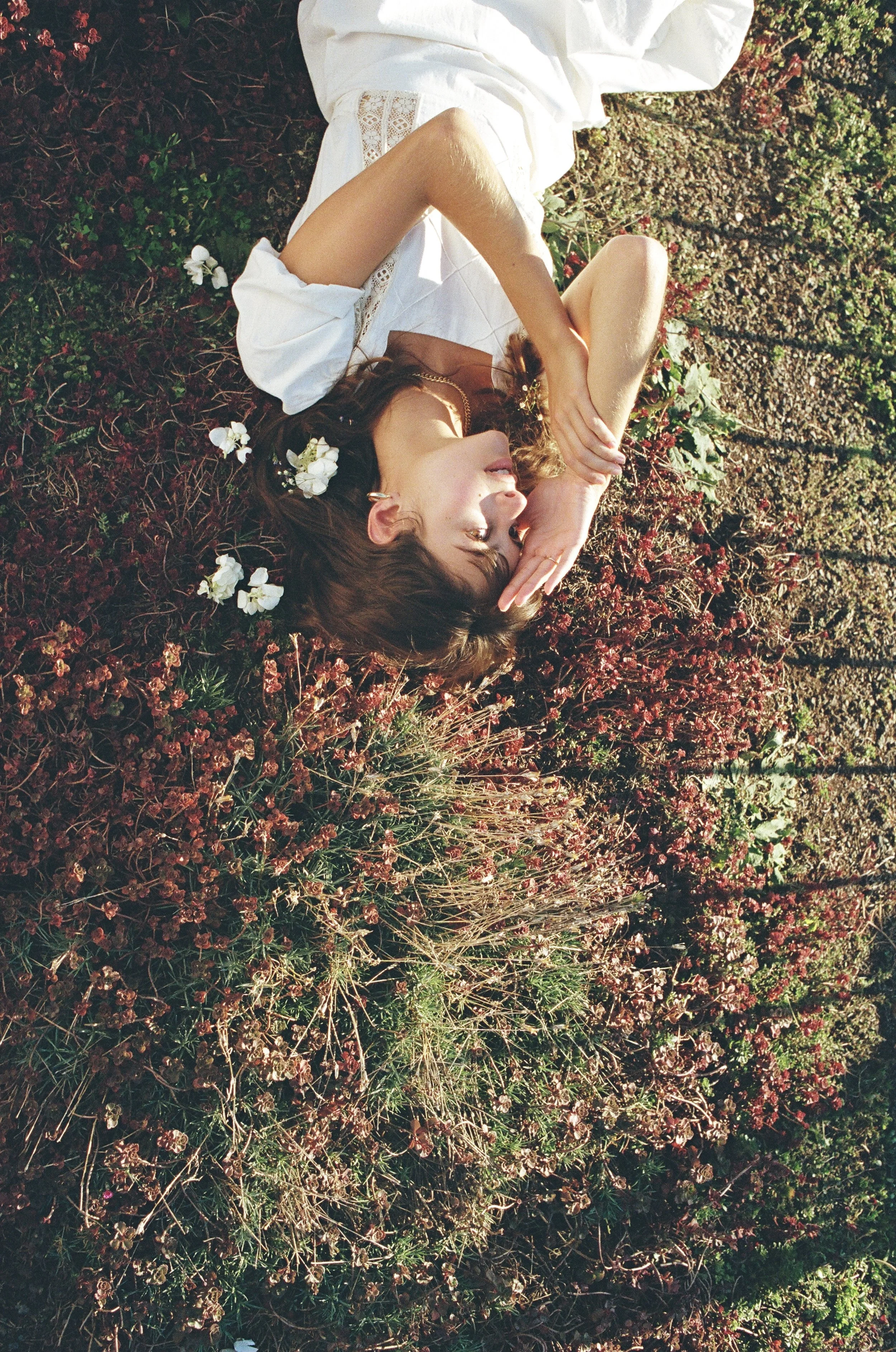 A woman lies on the ground amidst wildflowers and plants with her eyes closed, one hand covering part of her face, wearing a white dress with lace details.
