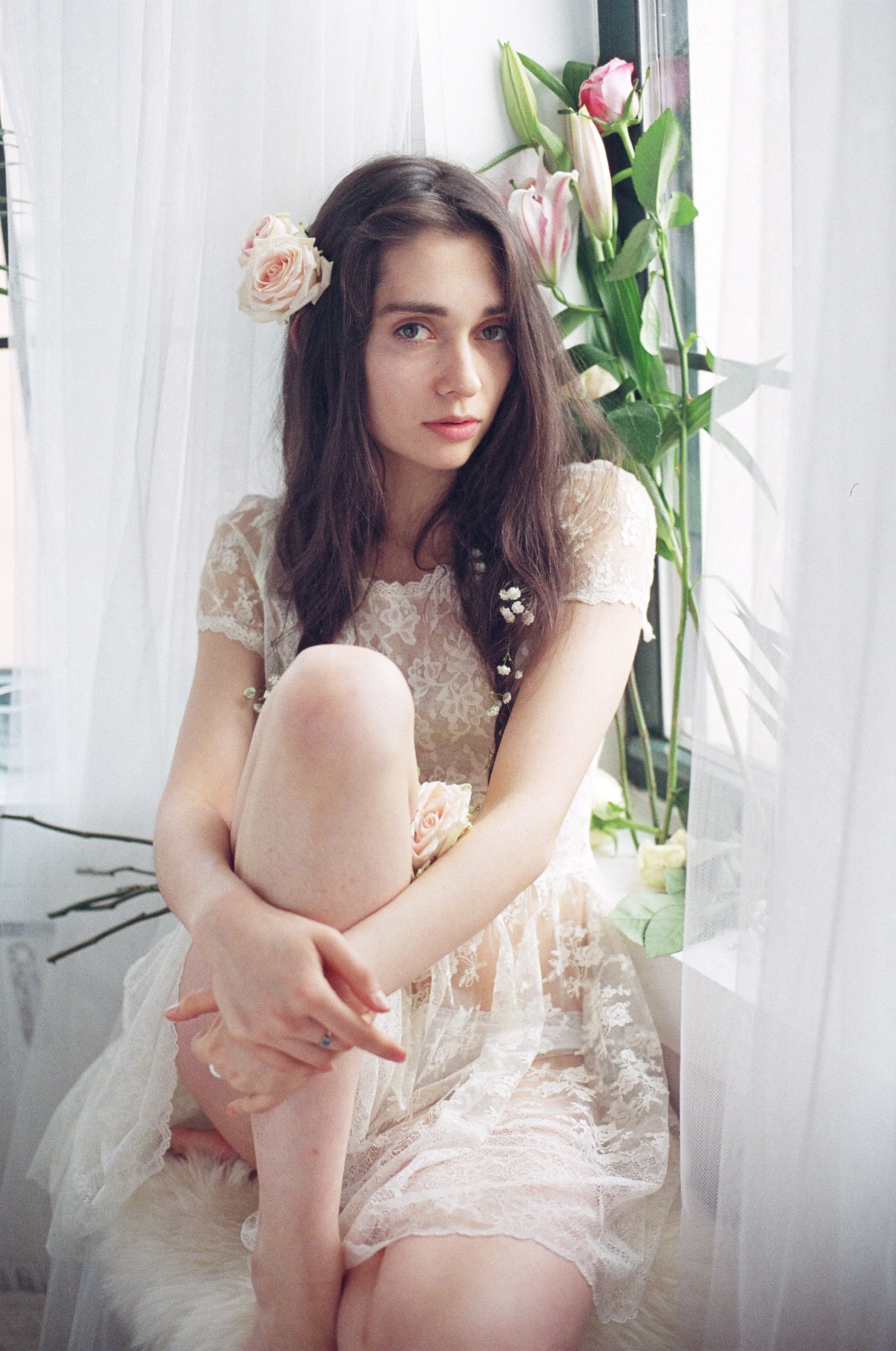 Young woman with long brown hair sitting by a window with pink flowers. She is wearing a lace dress and has a pink rose in her hair, with a contemplative expression.