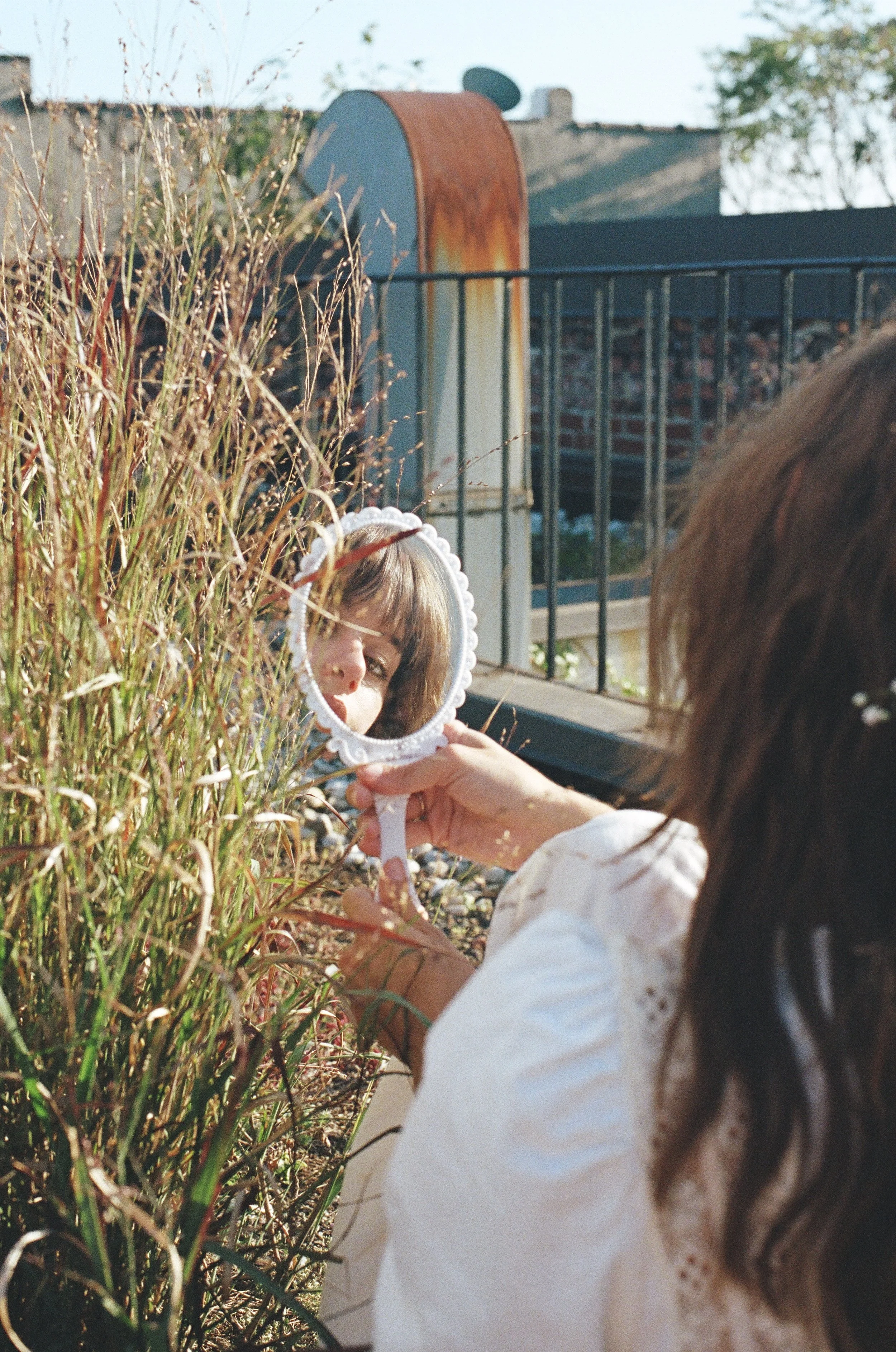 A woman with brown hair looking into a small round mirror with a white, decorative frame held in her hands. She is outdoors amid tall, dry grass, with a rusted cylindrical object and a fence in the background.