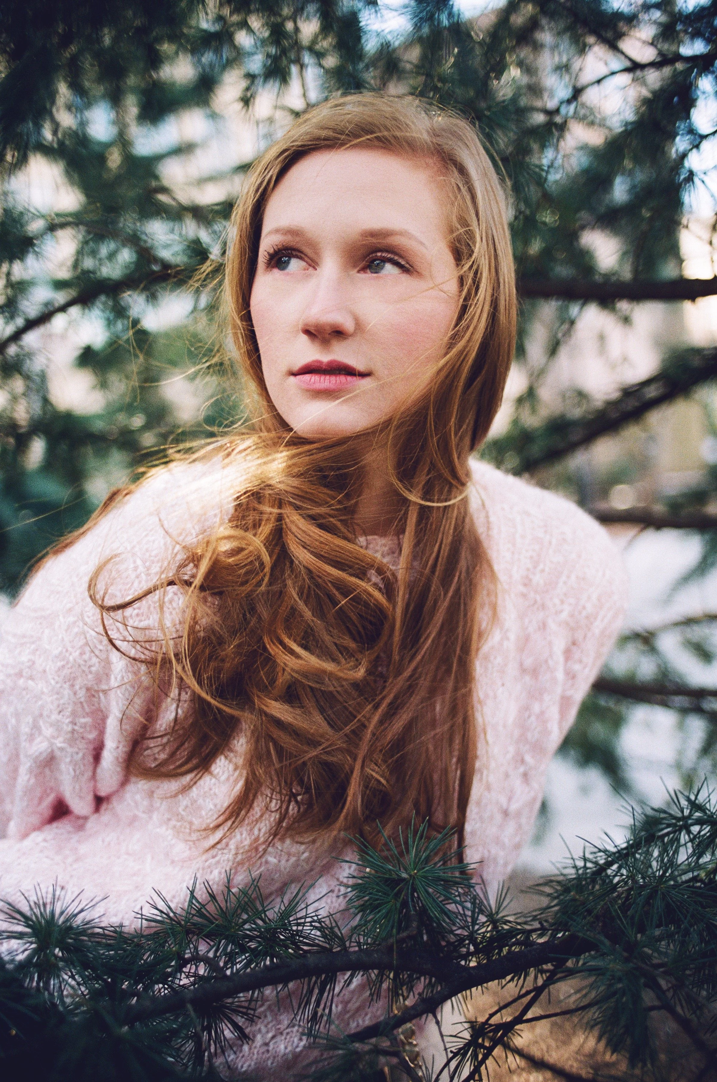 A woman with long, wavy red hair looking to her left, outdoors among pine trees.