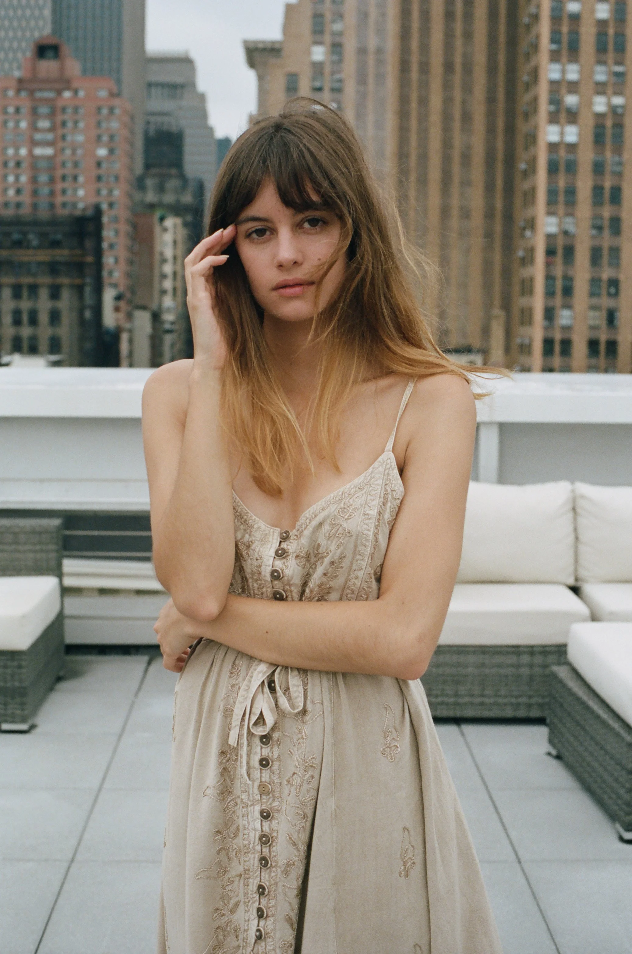 A young woman with long, wavy, brown hair, dressed in a beige summer dress, standing on a rooftop with city buildings in the background.
