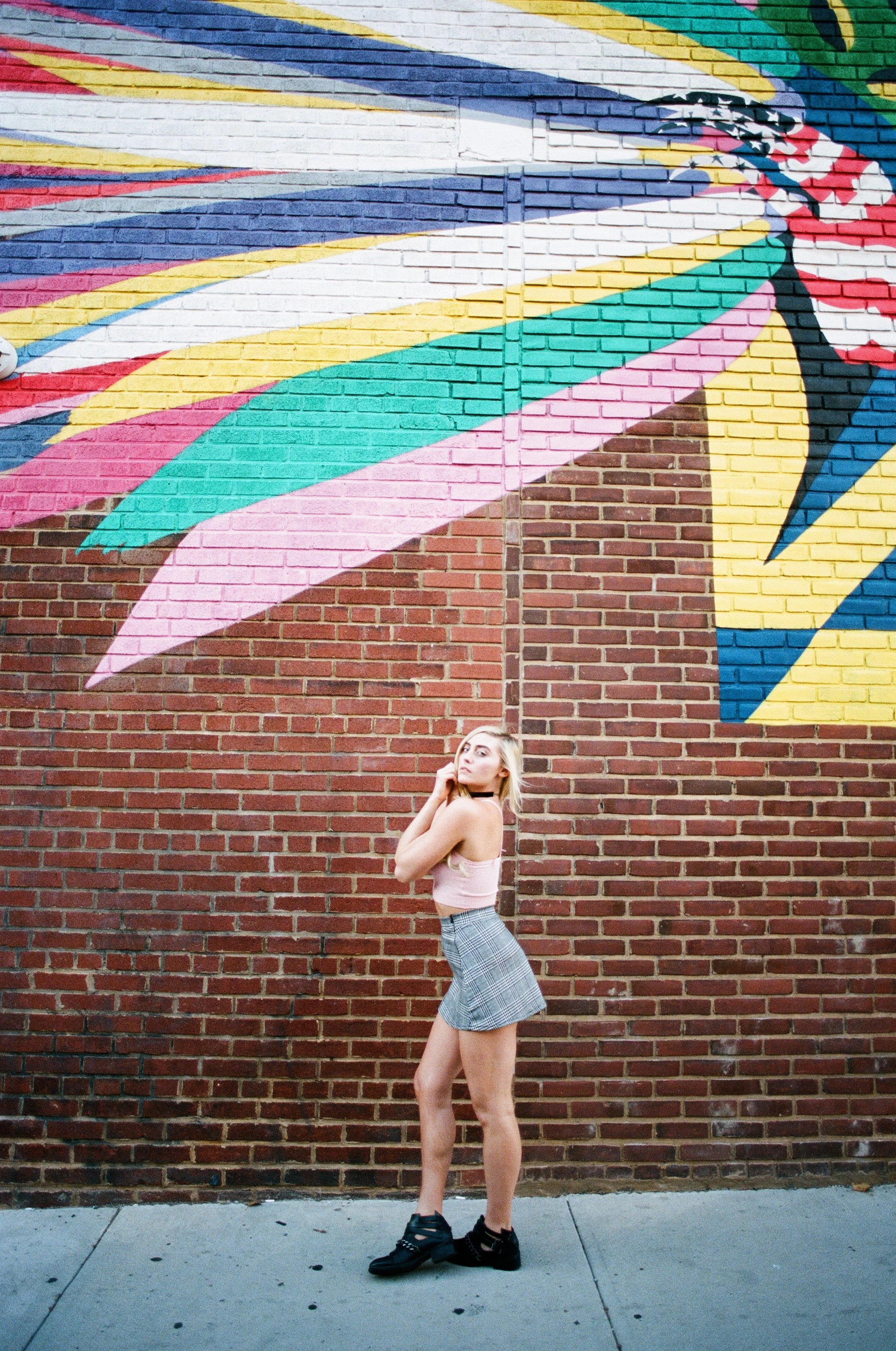 Young woman standing in front of a brick wall with colorful mural art, wearing a pink crop top, plaid skirt, black shoes, and a choker, posing with one hand on her neck and the other at her side.