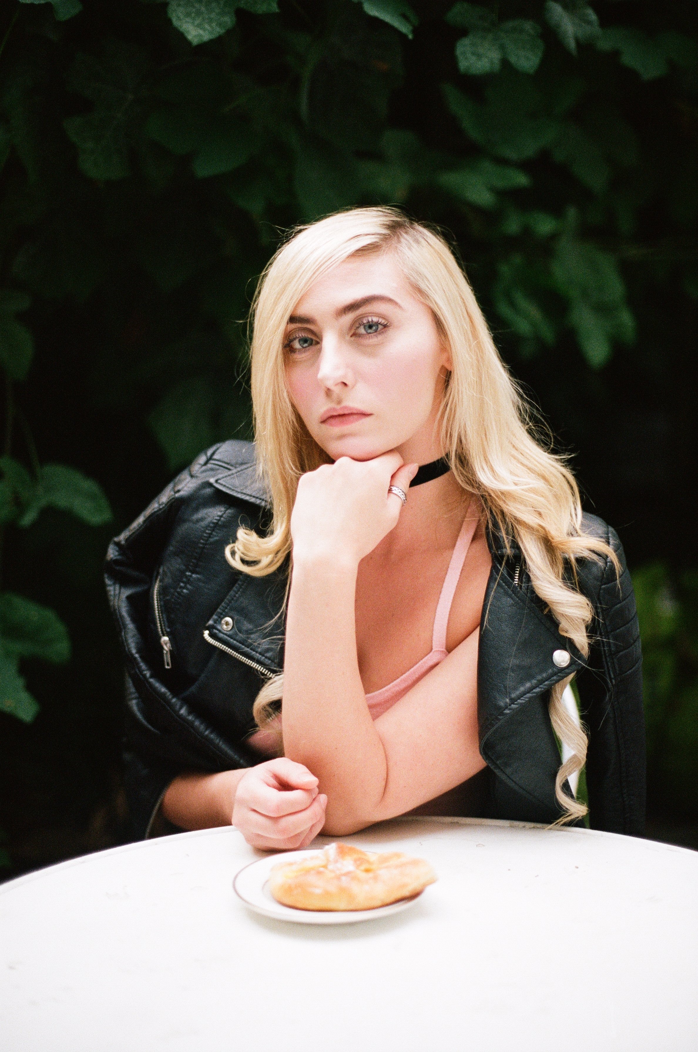 A young woman with blonde hair and fair skin sitting at a white table with a plate of food in front of her, wearing a black leather jacket, light pink top, and a black choker, with greenery in the background.