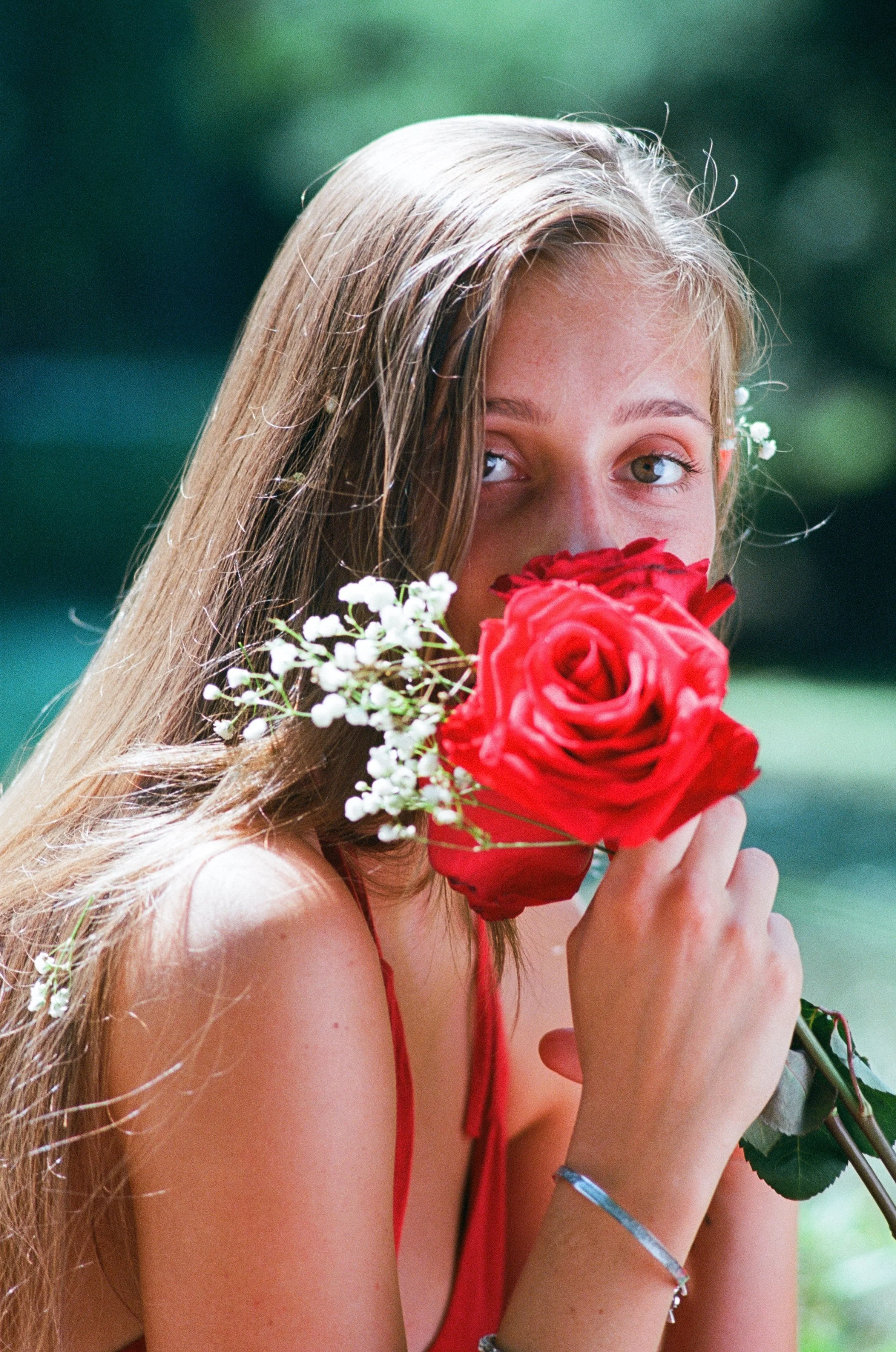 Young woman with long hair holding a bouquet of red roses and white flowers near her face, outdoors with blurred green background.