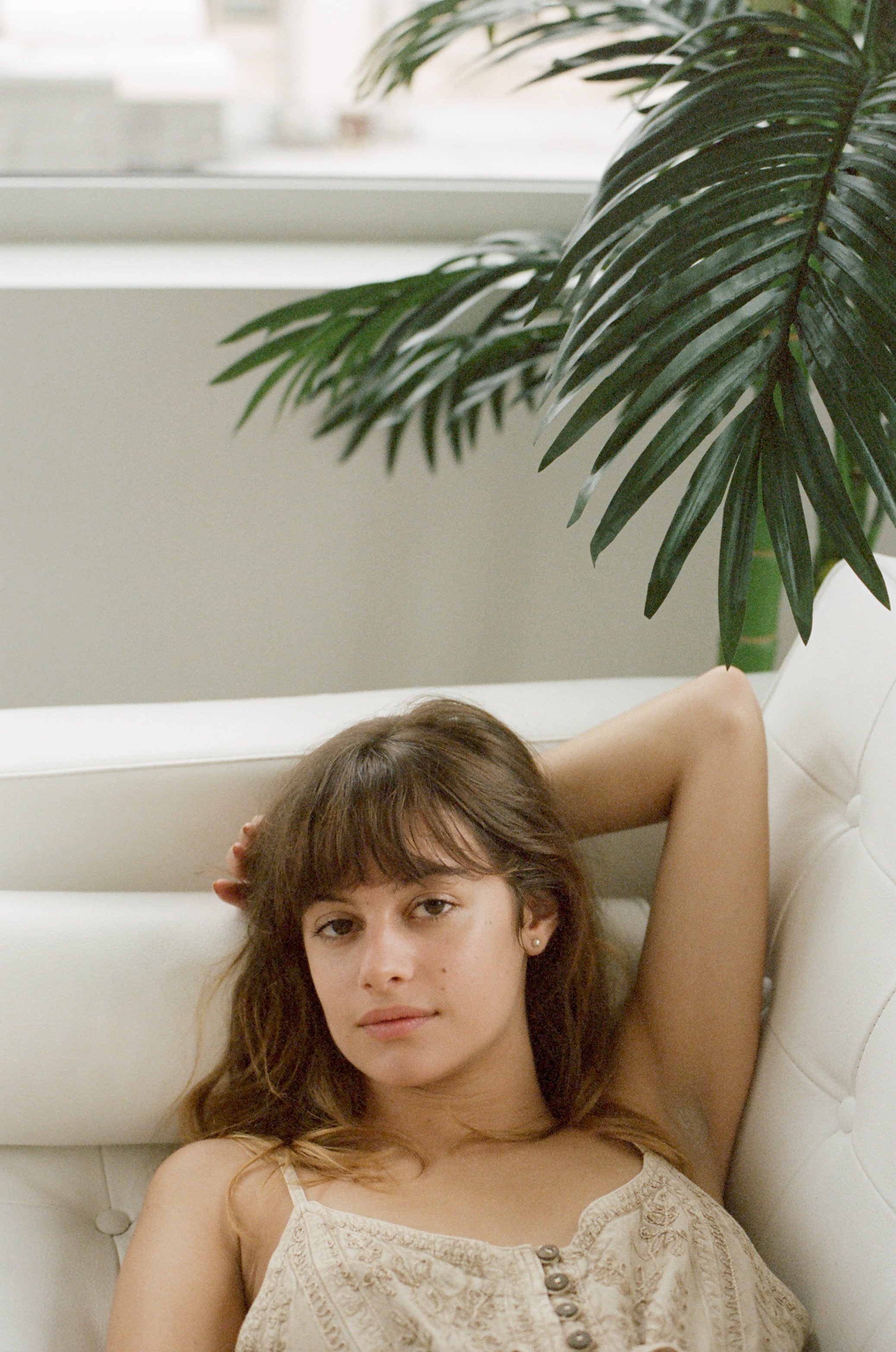 A woman with brown hair and a beige top relaxing on a white leather chair next to a large green indoor plant, near a window.