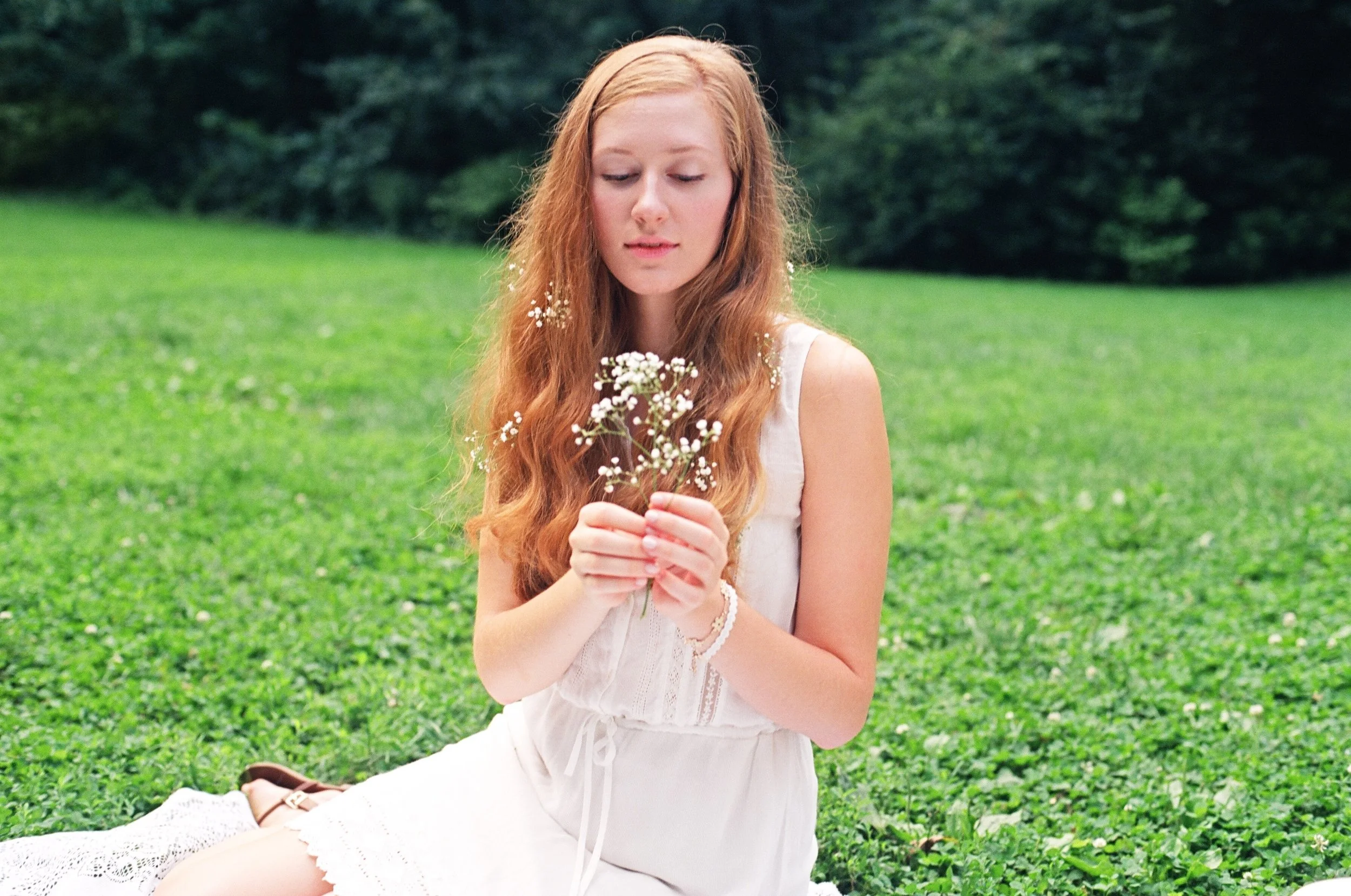 A young woman with long red hair sits on a grassy field, holding a small bouquet of white flowers, wearing a white sleeveless dress, and surrounded by greenery.