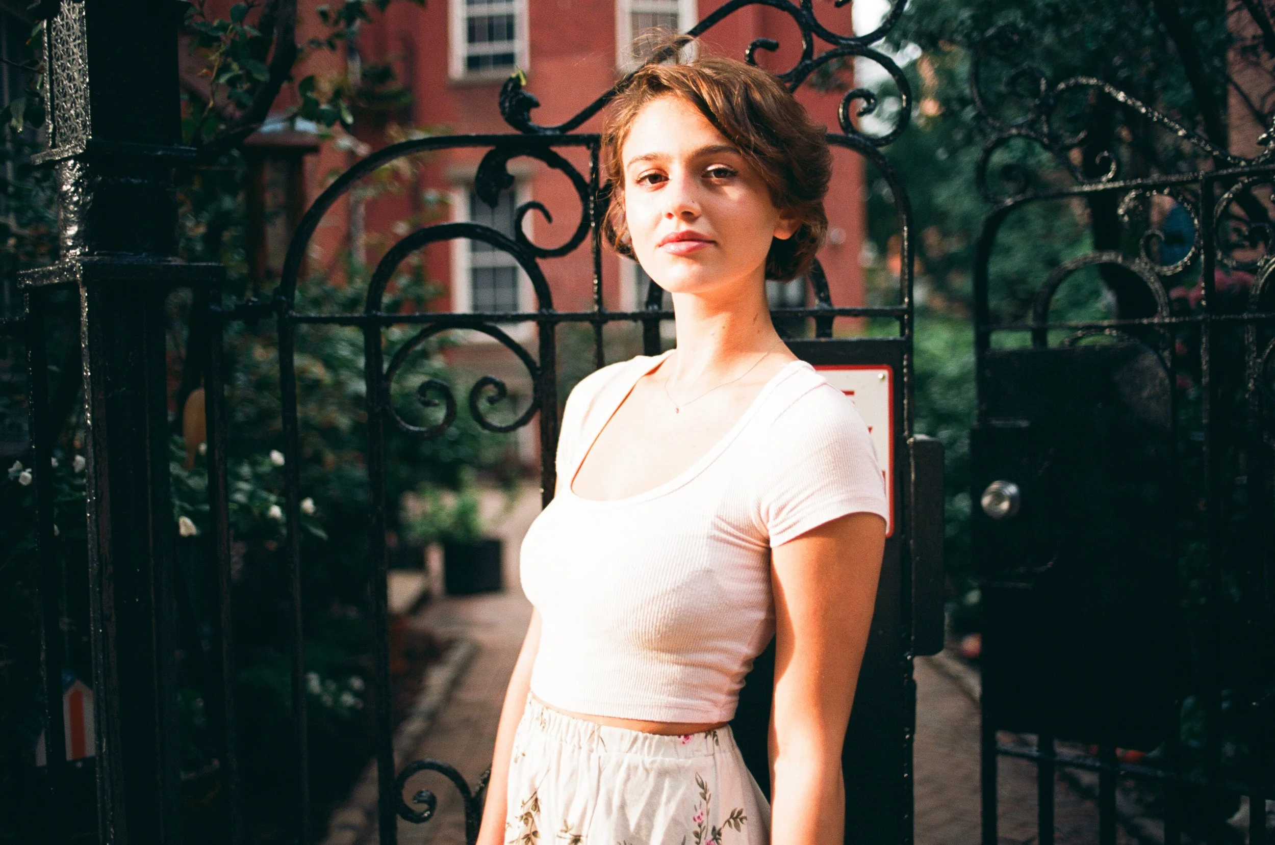 A young woman with short brown hair, wearing a white crop top and floral skirt, standing in front of a black iron gate with a red building in the background.