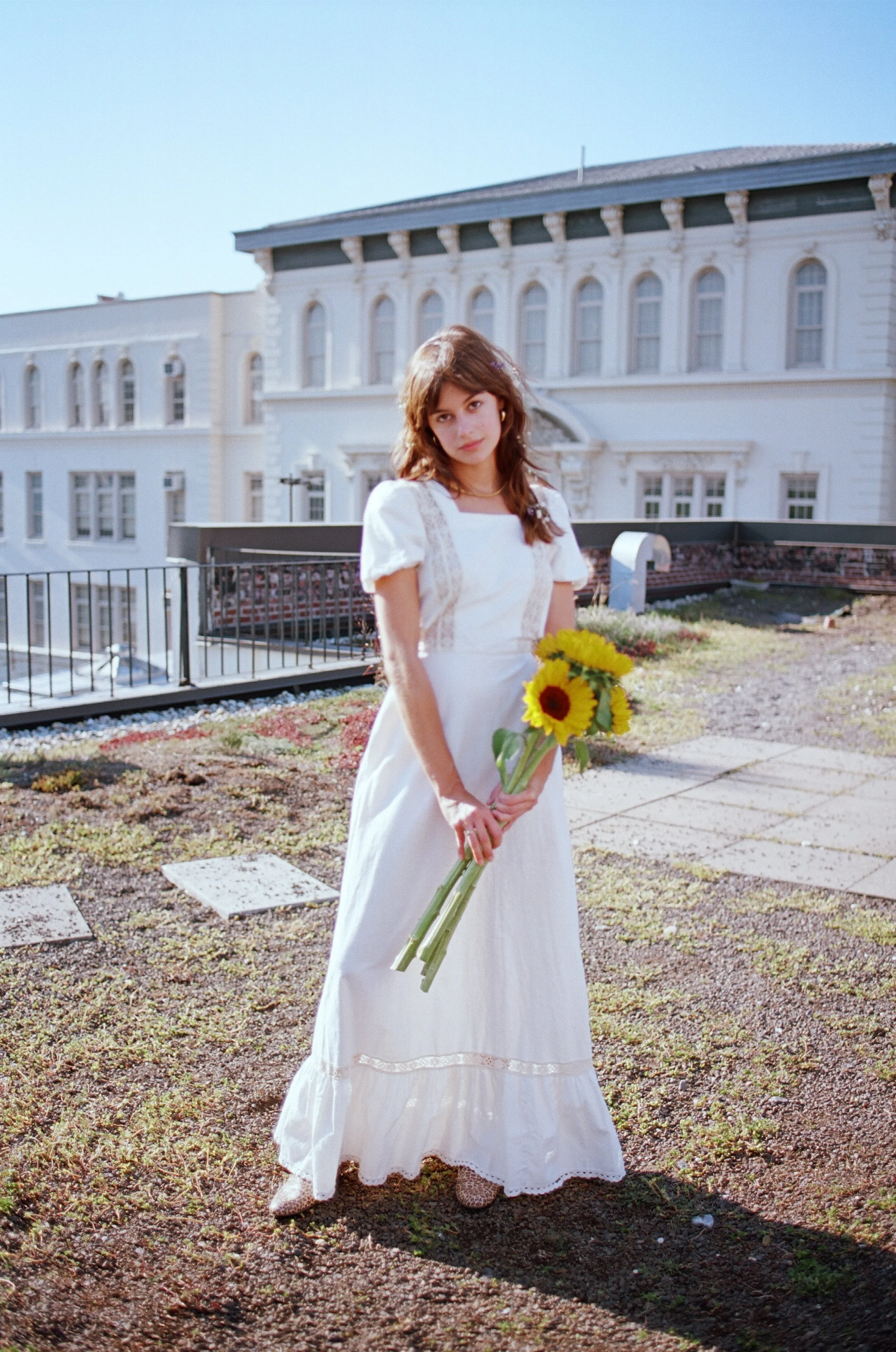 A young woman in a white dress holding a bouquet of sunflowers on a rooftop.