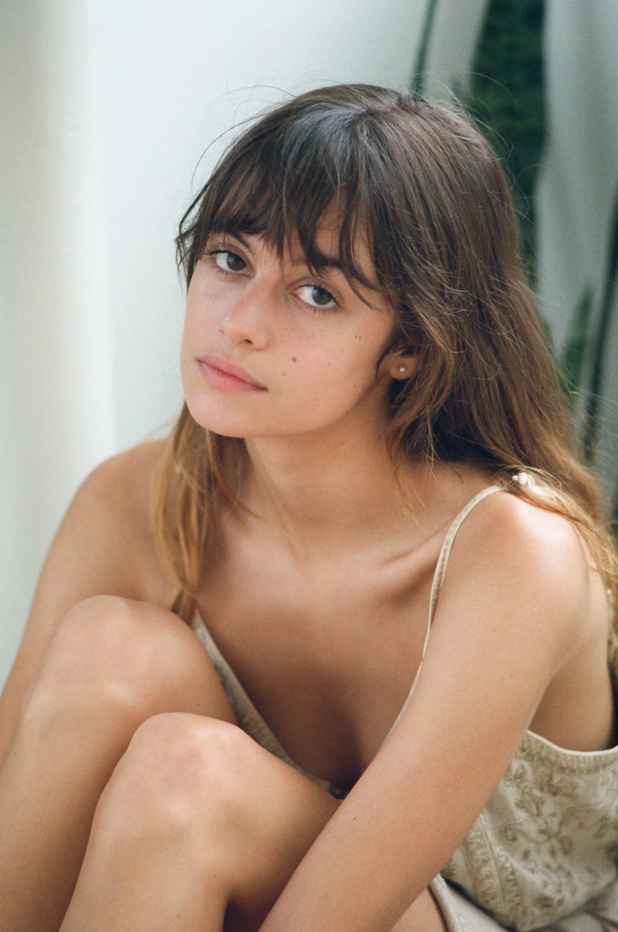 A young woman with long, wavy brown hair and blue eyes, sitting with her arms around her legs, wearing a beige camisole, looking directly at the camera.