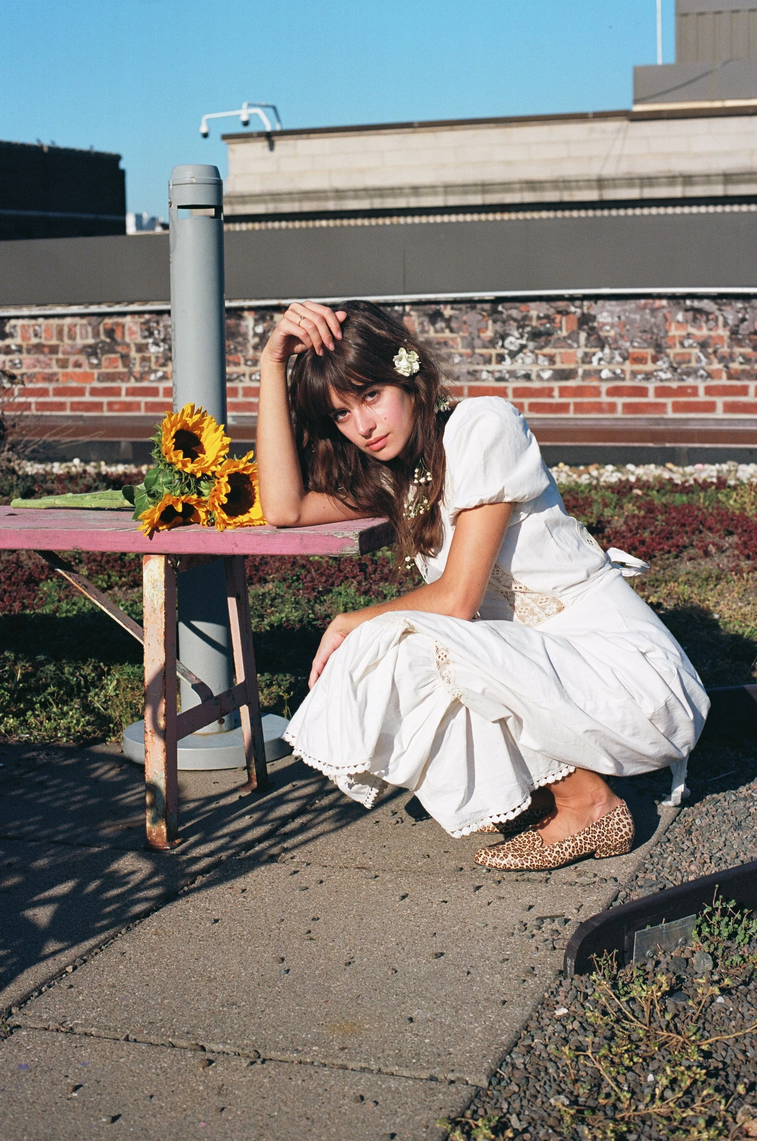 A young woman with brown hair, wearing a white dress and leopard print flats, sits on the sidewalk next to a pink table with a bouquet of sunflowers, resting her head on her hand with a cityscape in the background.