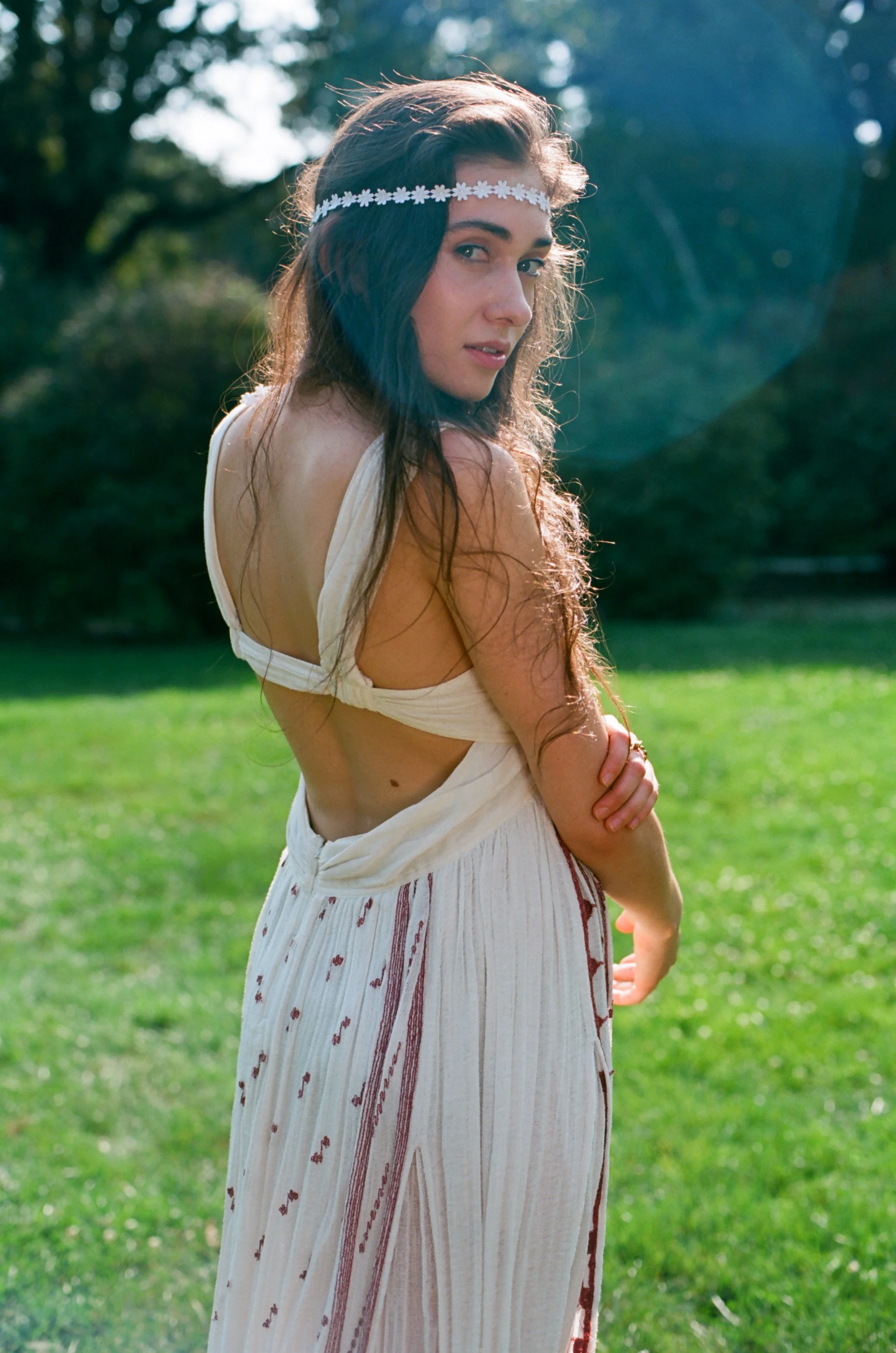 A young woman with long dark hair wearing a white flower headband and a cream-colored dress with cutouts stands outdoors on a green lawn with trees in the background.