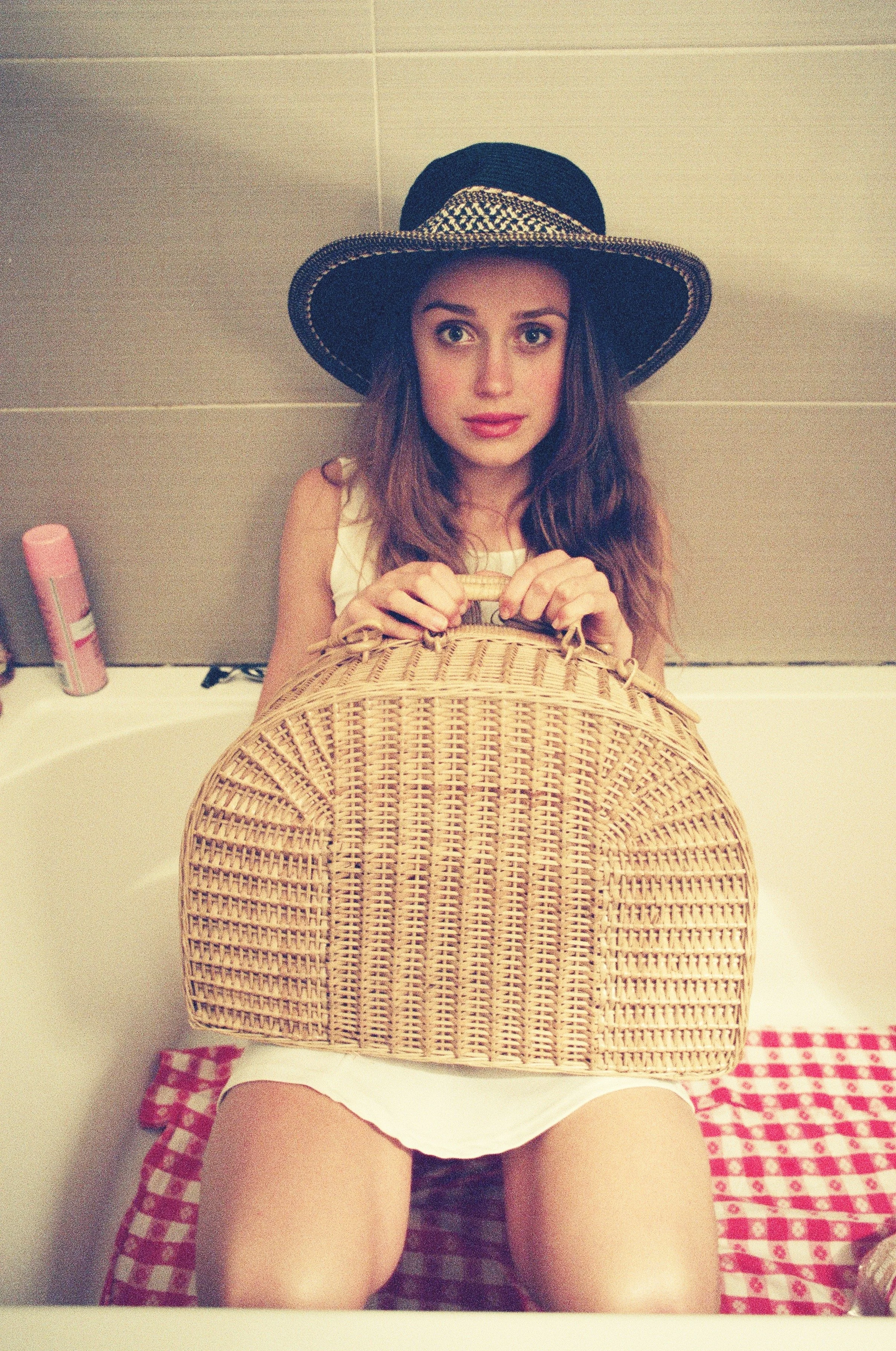 A young woman with brown hair wearing a wide-brimmed hat, sitting in a bathtub, holding a woven basket with both hands, and looking at the camera. Photo edited by Mary Perrino.