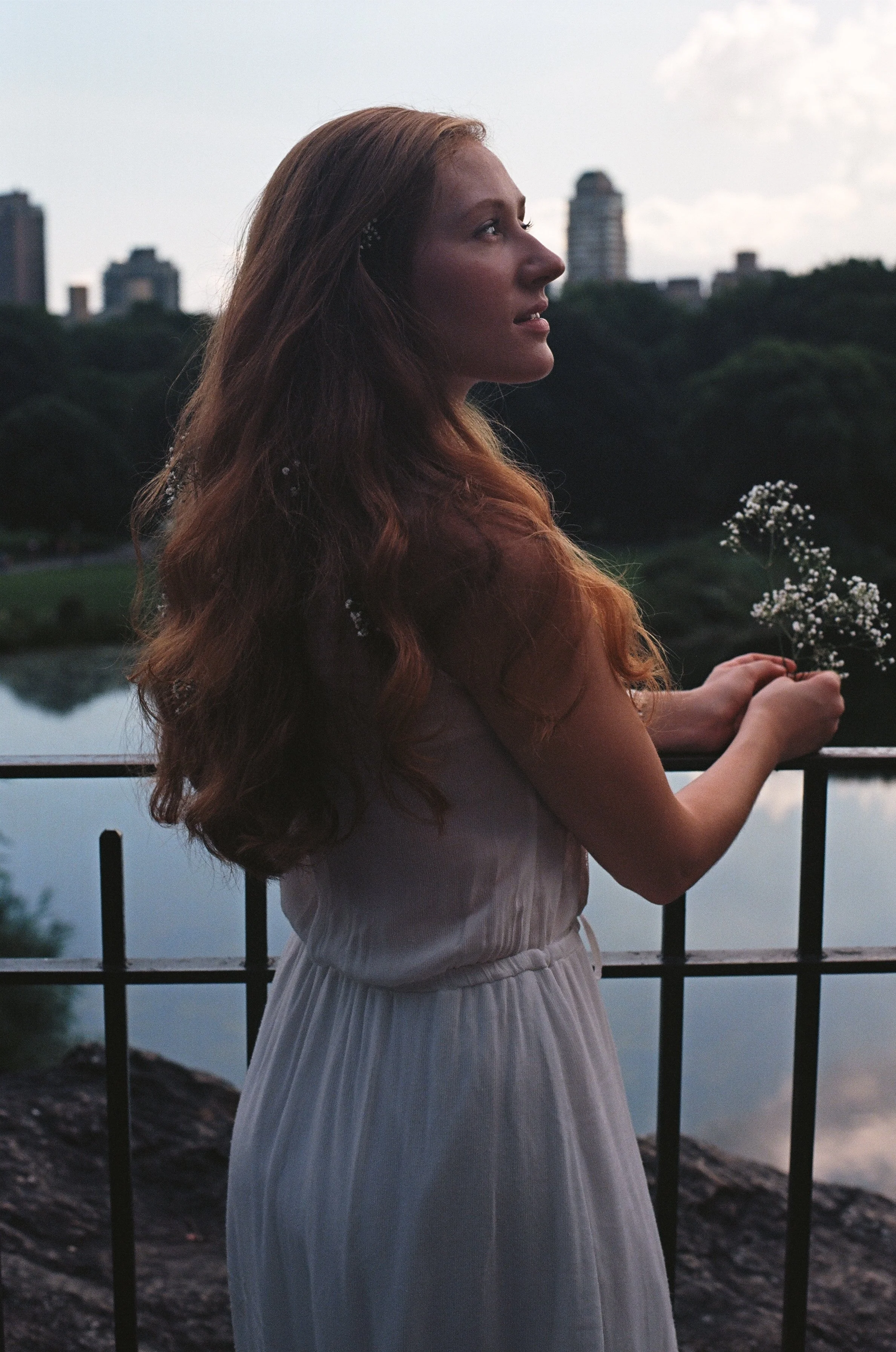 A woman with long red hair in a white dress standing by a railing near a body of water, holding a small bouquet of white flowers, with city buildings and trees in the background during dusk.