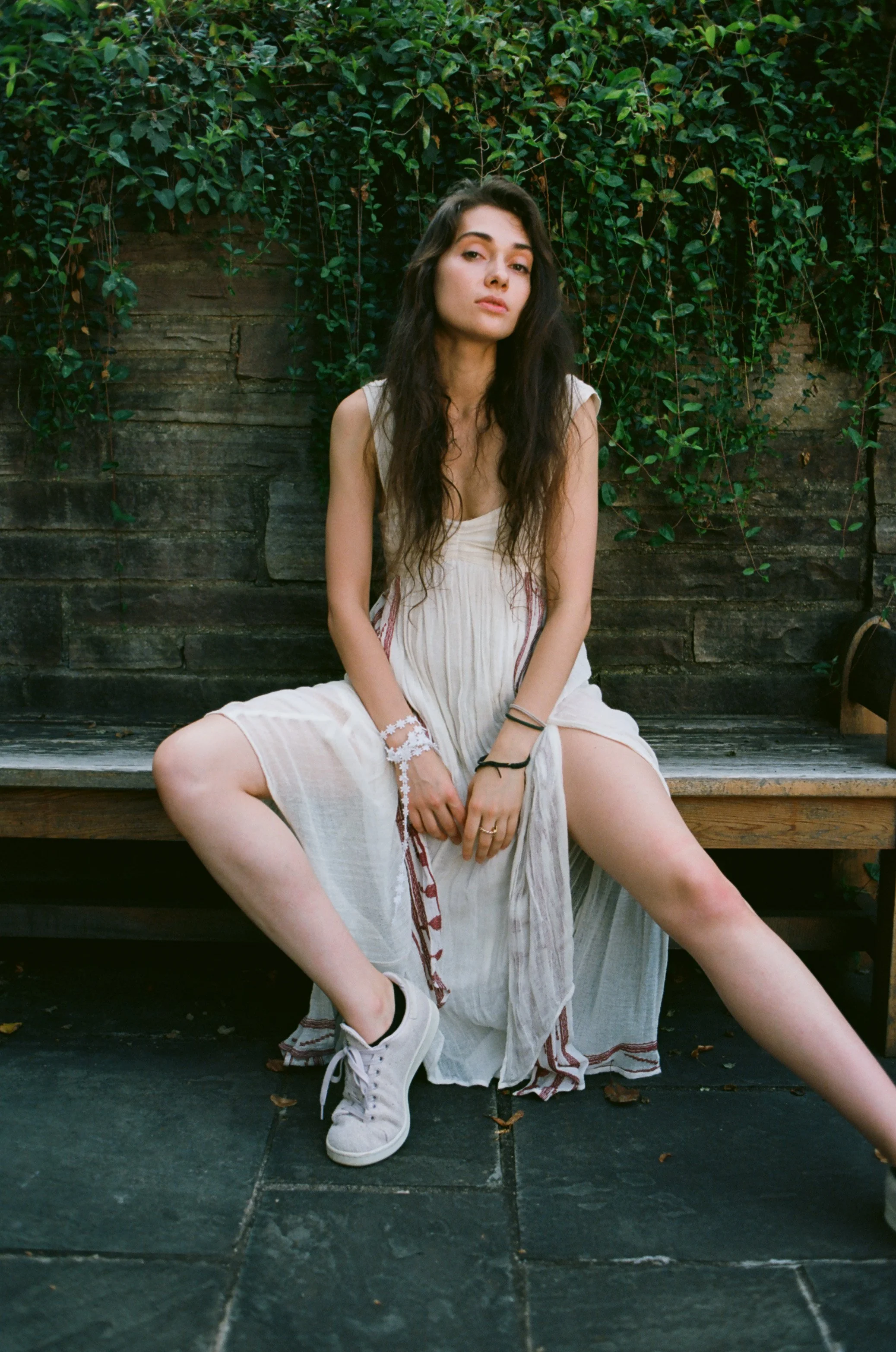 A young woman with long dark hair, wearing a light-colored dress and white sneakers, sitting on a wooden bench in front of a stone wall with green vines. Photo edited by Mary Perrino.