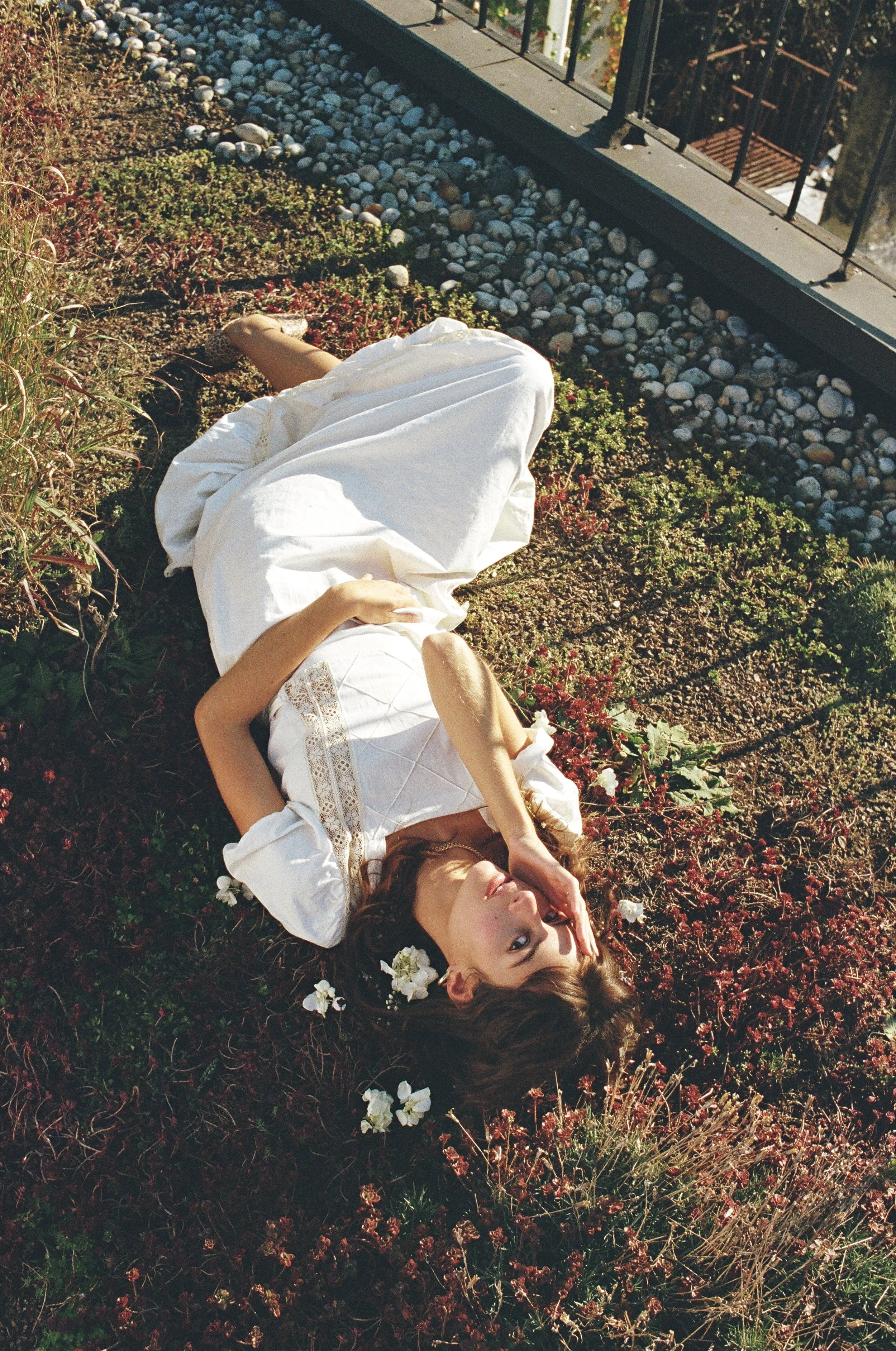 A woman in a white dress with flowers in her hair lying on garden soil surrounded by plants, with a fence and gravel area nearby, basking in sunlight.
