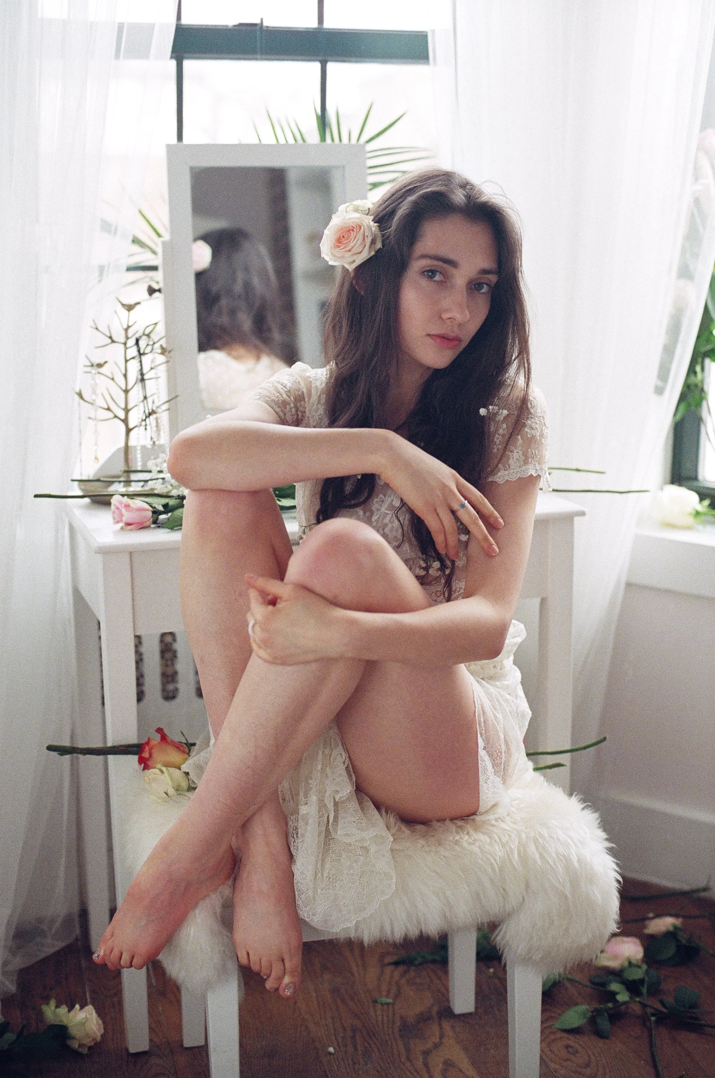 Young woman with long dark hair and a flower in her hair, sitting on a fluffy stool in a bright room, surrounded by roses and decorative plants, gazing at the camera with a relaxed expression.