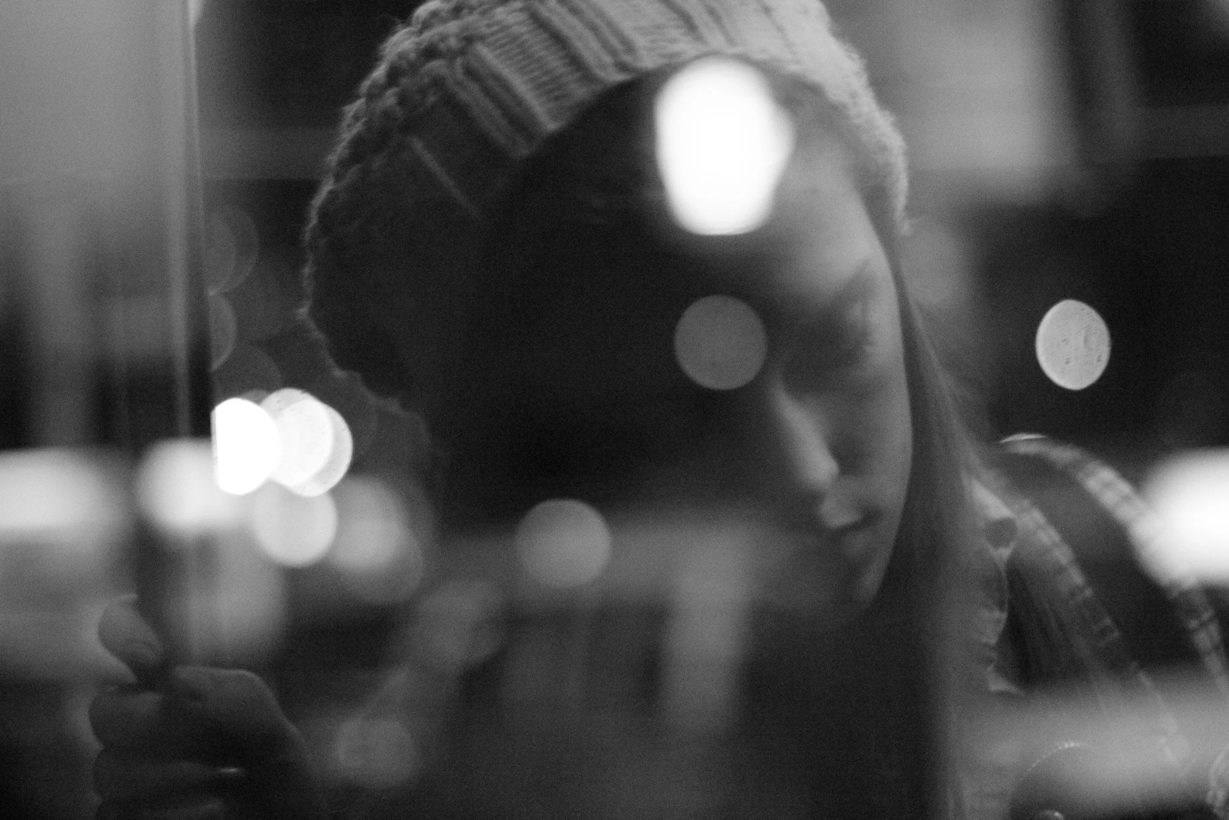 A black and white photo of a young girl with long hair, wearing a knitted beanie, looking down with a thoughtful expression. The background includes blurred city lights. Photo edited by Mary Perrino.