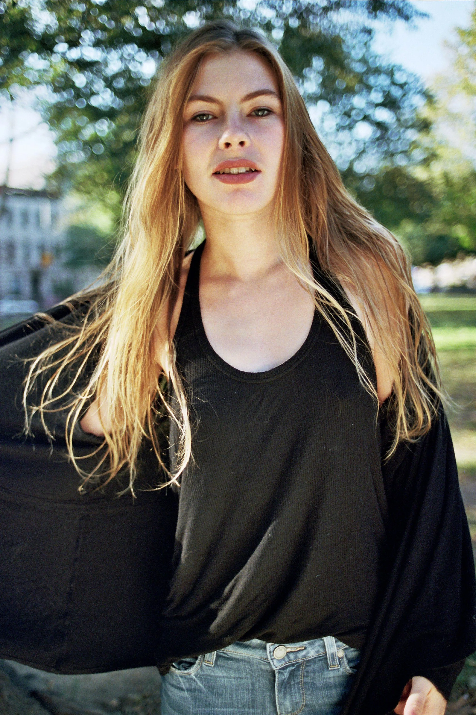 A young woman with long, wavy blonde hair wearing a black tank top and jeans outdoors on a sunny day with trees and houses in the background.