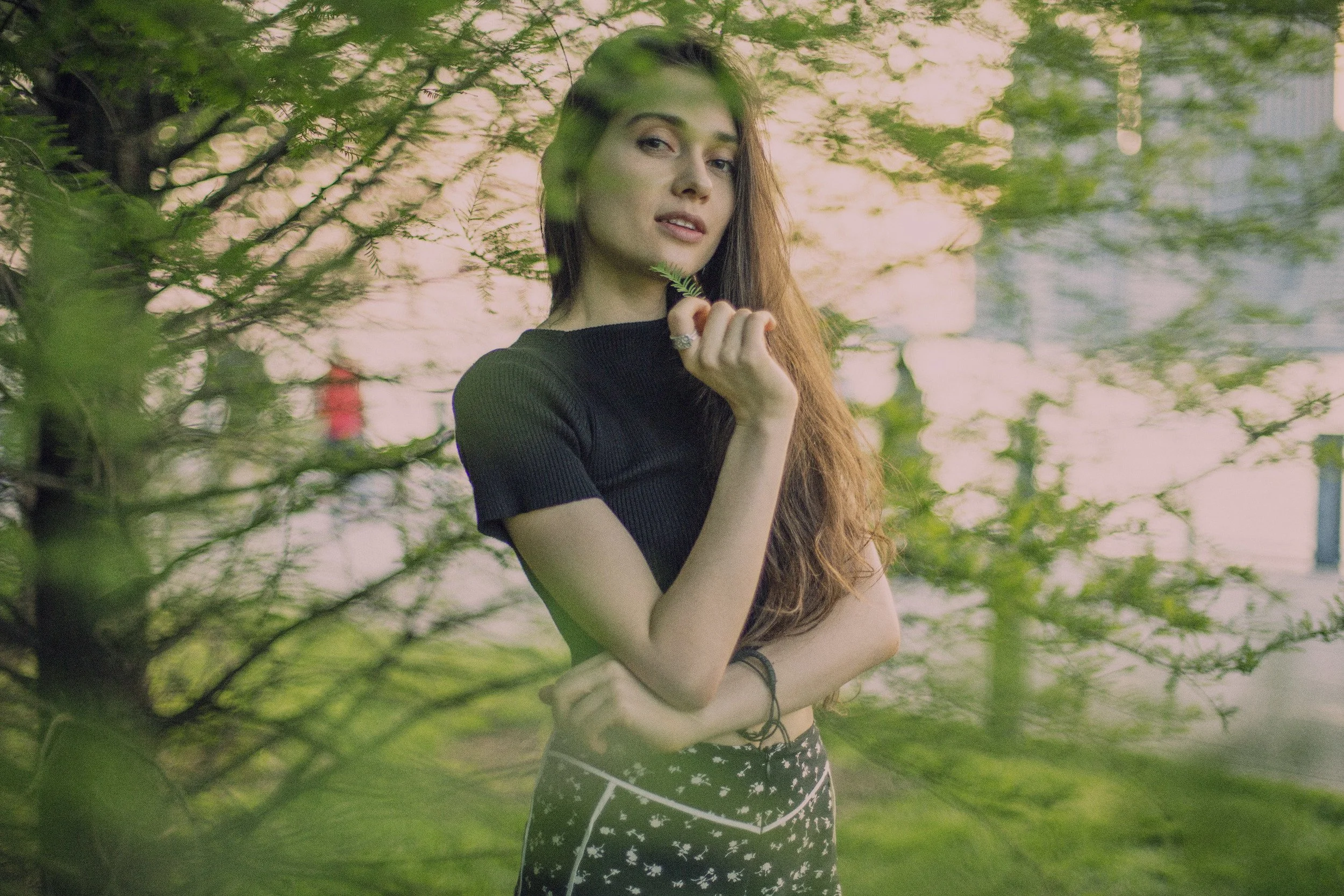 Young woman with long, wavy hair, wearing a black top and patterned pants, standing outdoors among green trees, holding a small branch and looking at the camera.
