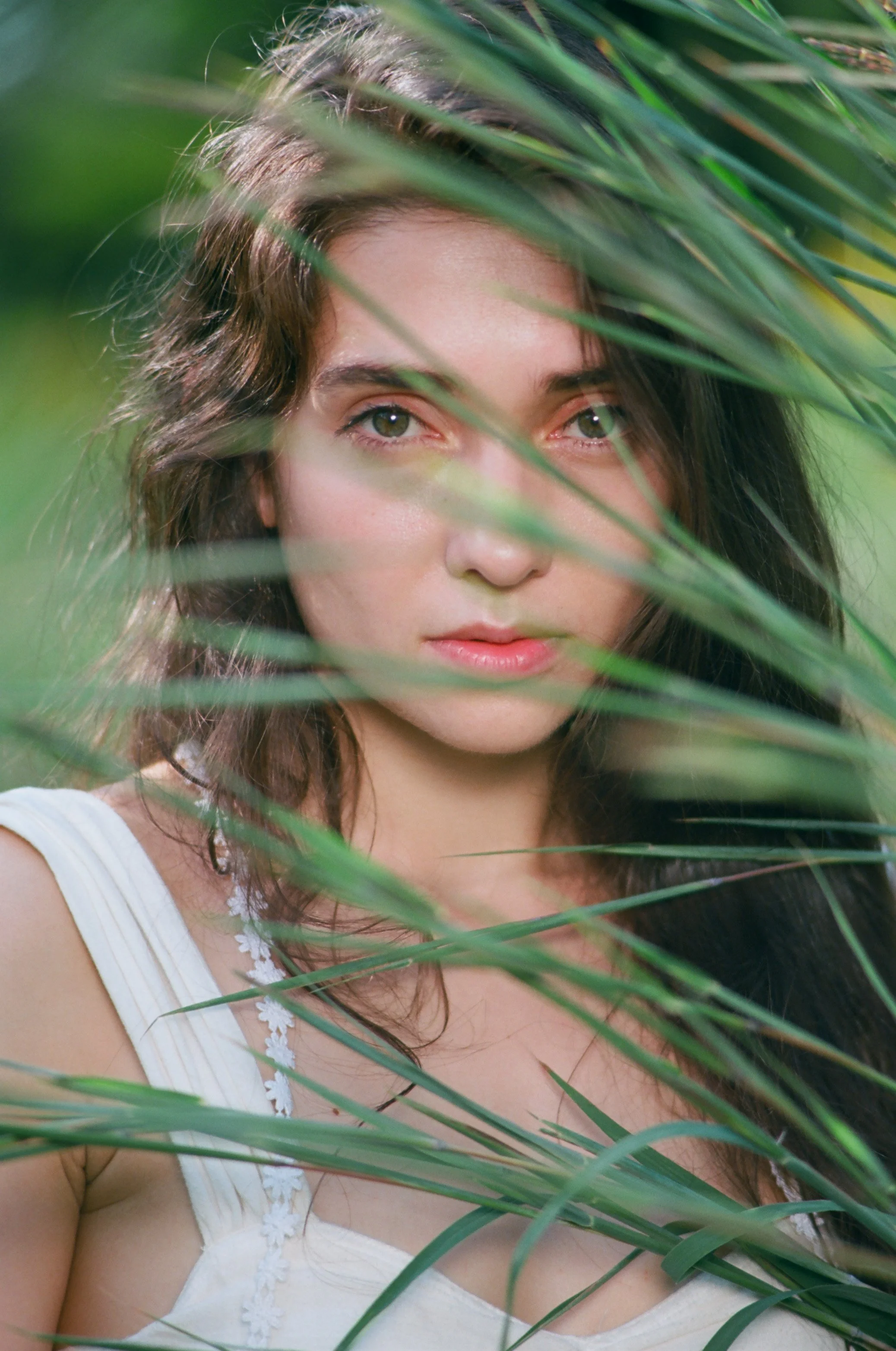 A young woman with brown hair and blue eyes is partially obscured by green palm leaves, with a blurred green background.