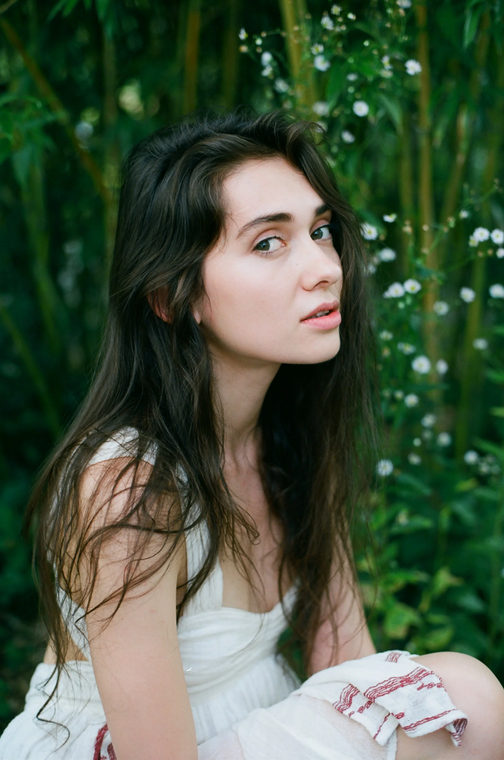 Young woman with long dark hair and blue eyes sitting outdoors in a natural setting with green plants and small white flowers.