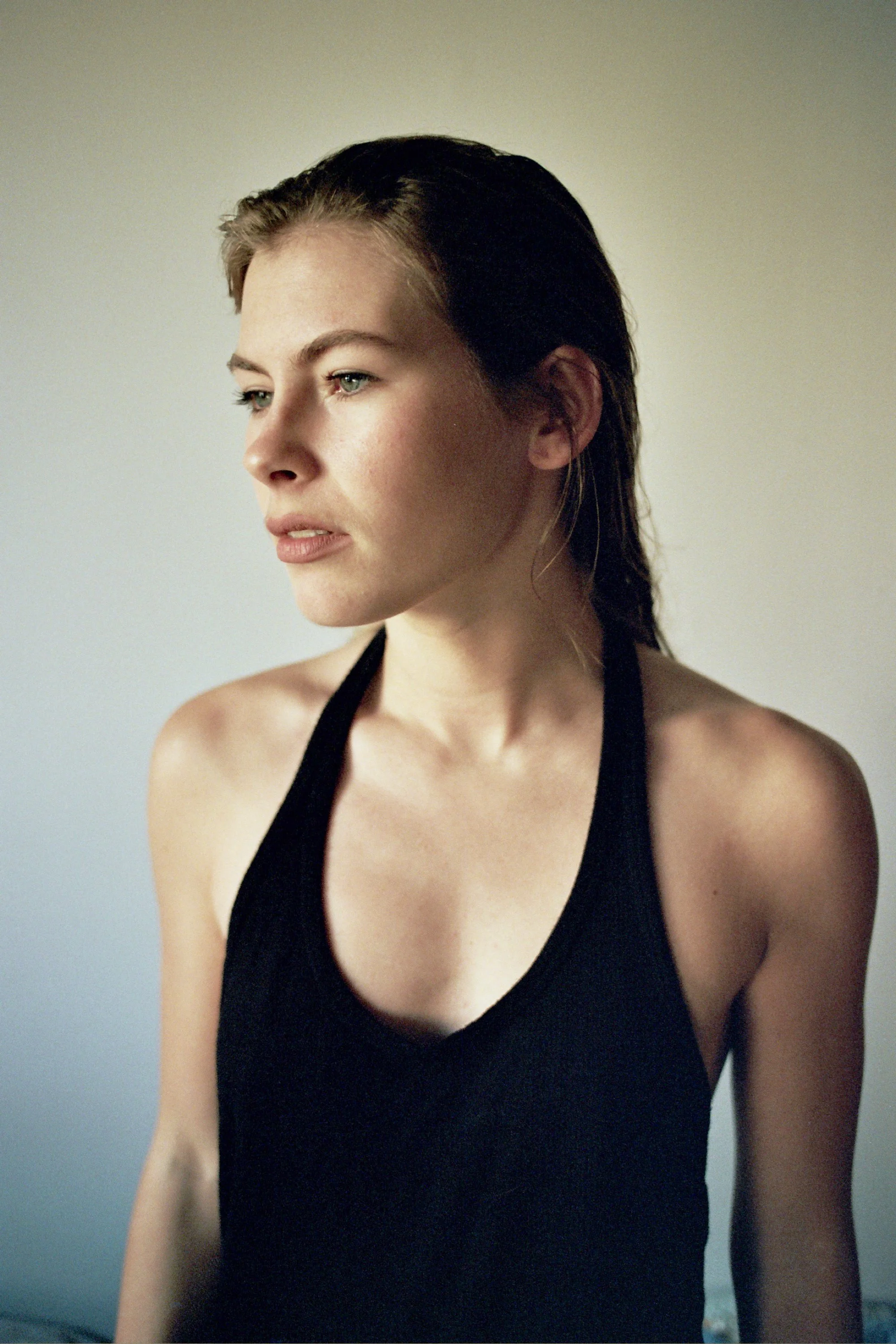 A young woman with light skin and dark, wet hair wearing a black sleeveless top, looking contemplative against a plain, light background. Photo edited by Mary Perrino.
