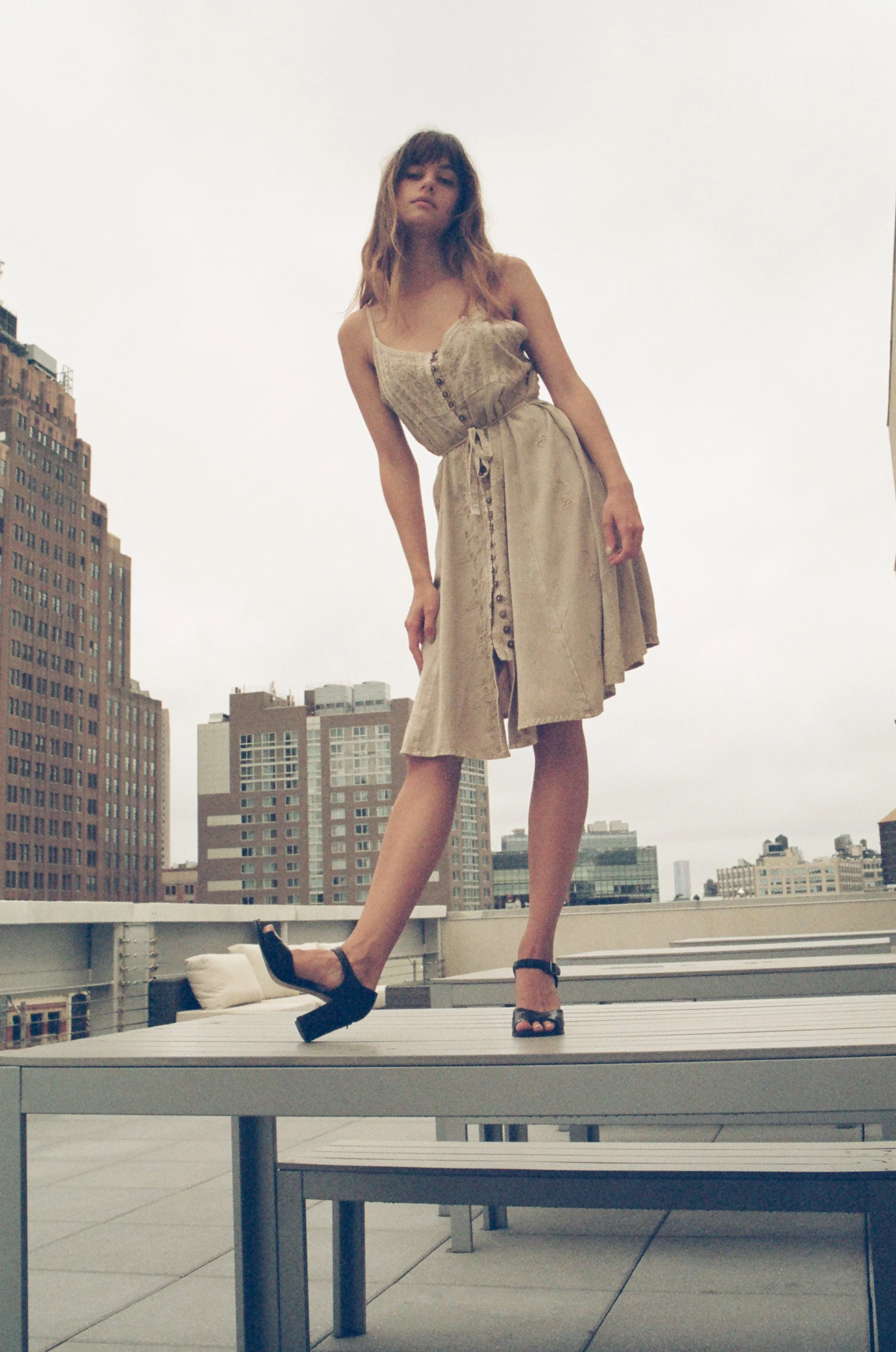 A woman in a beige dress and high heels standing on an outdoor rooftop with city skyscrapers in the background.