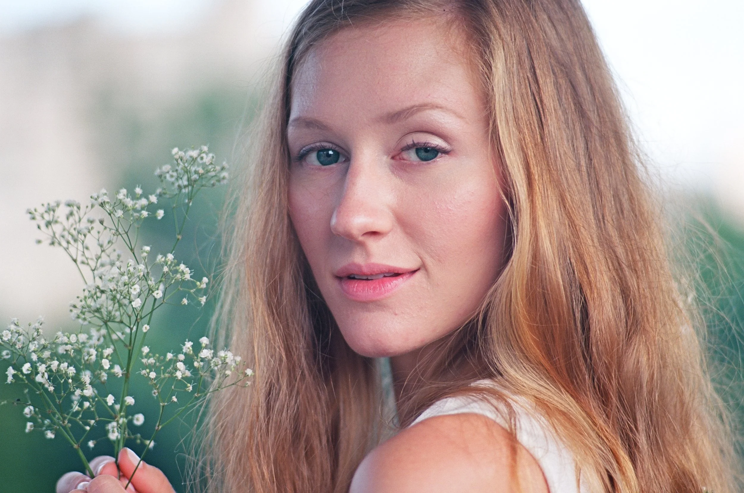 A woman with long red hair and blue eyes holding a small bunch of white flowers, standing outdoors with greenery in the background.