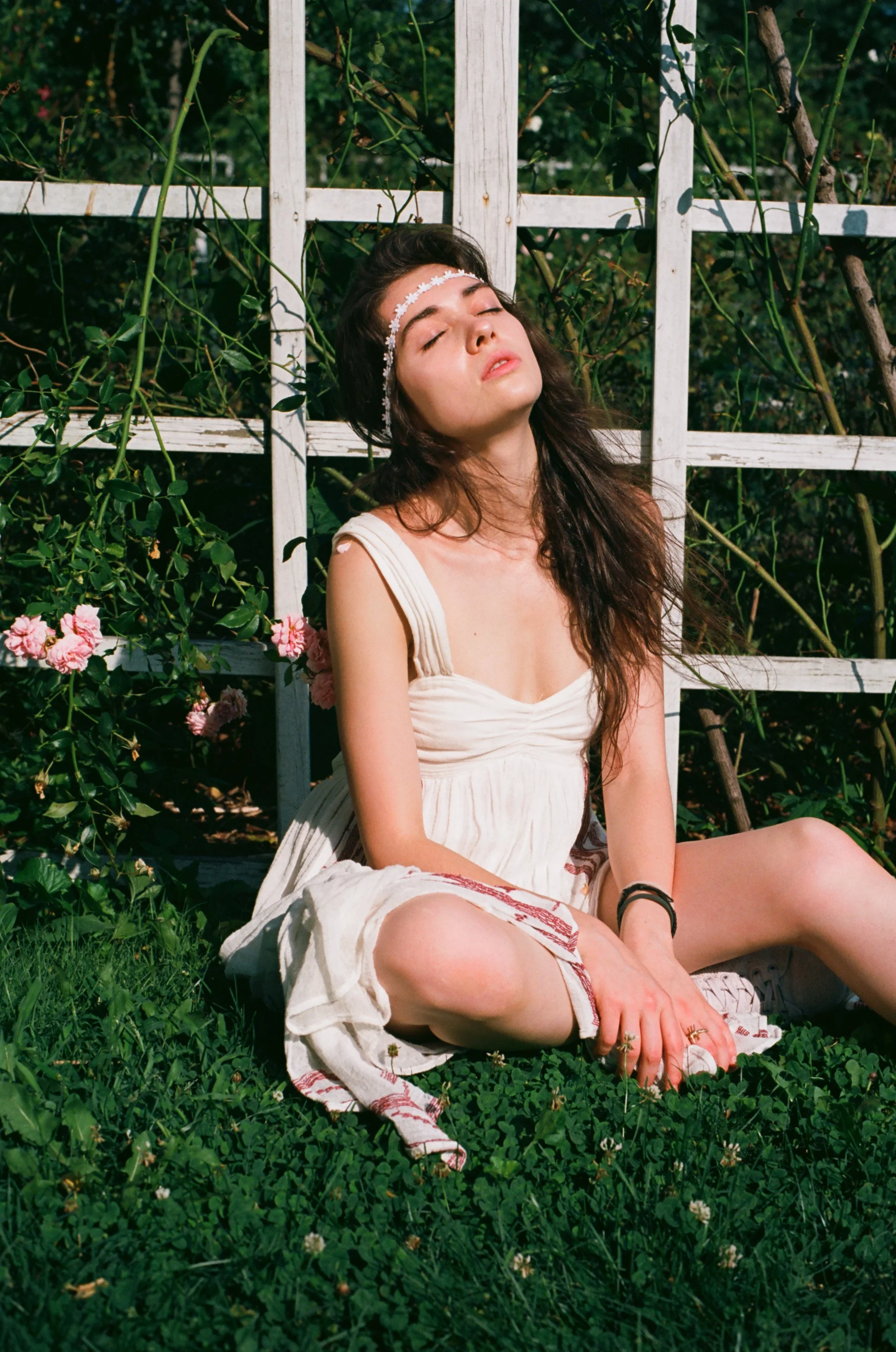 Young woman sitting on grass with eyes closed, wearing a white dress and a floral headband, surrounded by greenery and pink flowers.