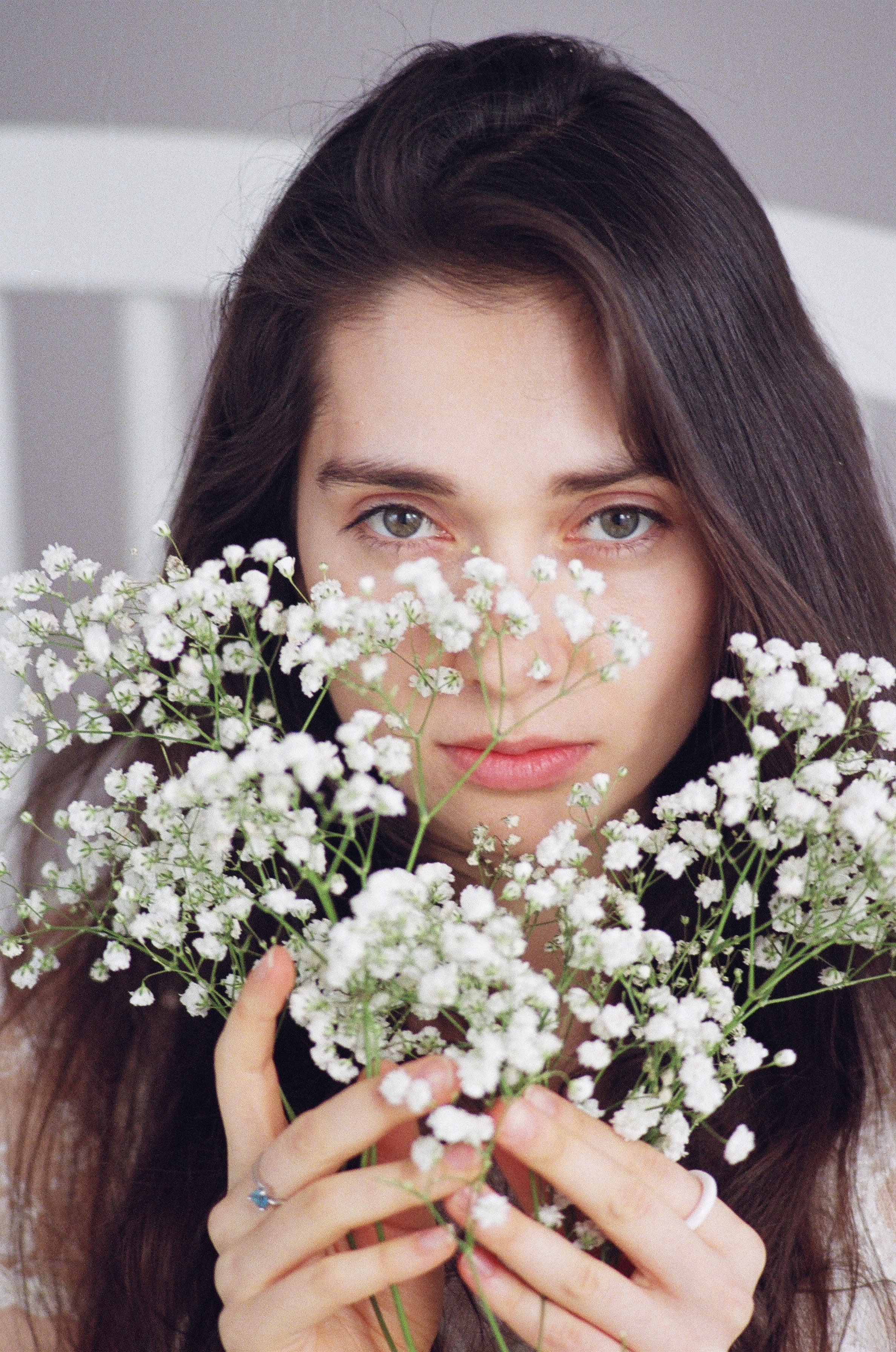 A young woman with long brown hair and light eyes holding a bouquet of white baby's breath flowers close to her face.