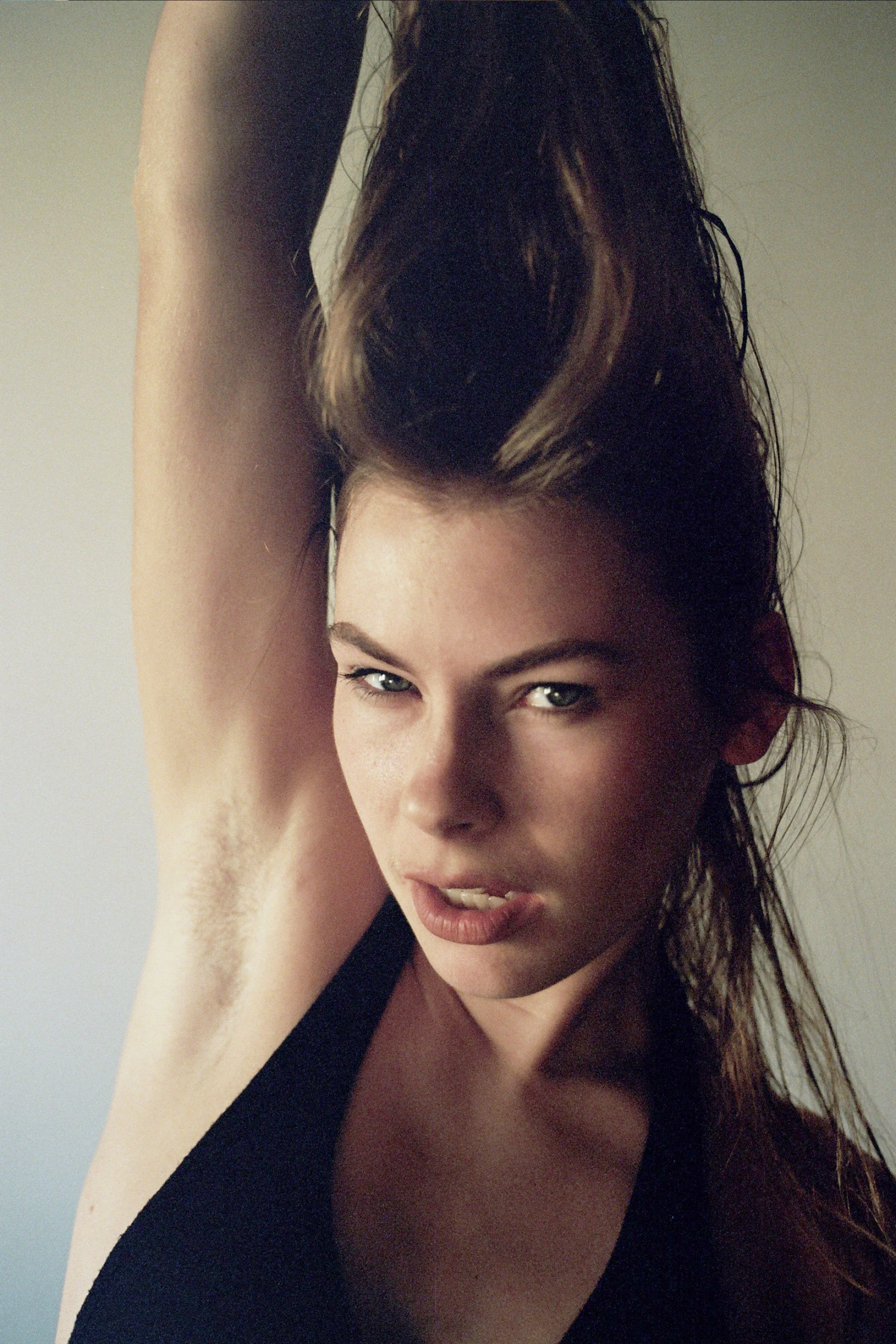 A woman with brown hair, blue eyes, and light skin is posing with her right arm raised above her head, showing her underarm.
