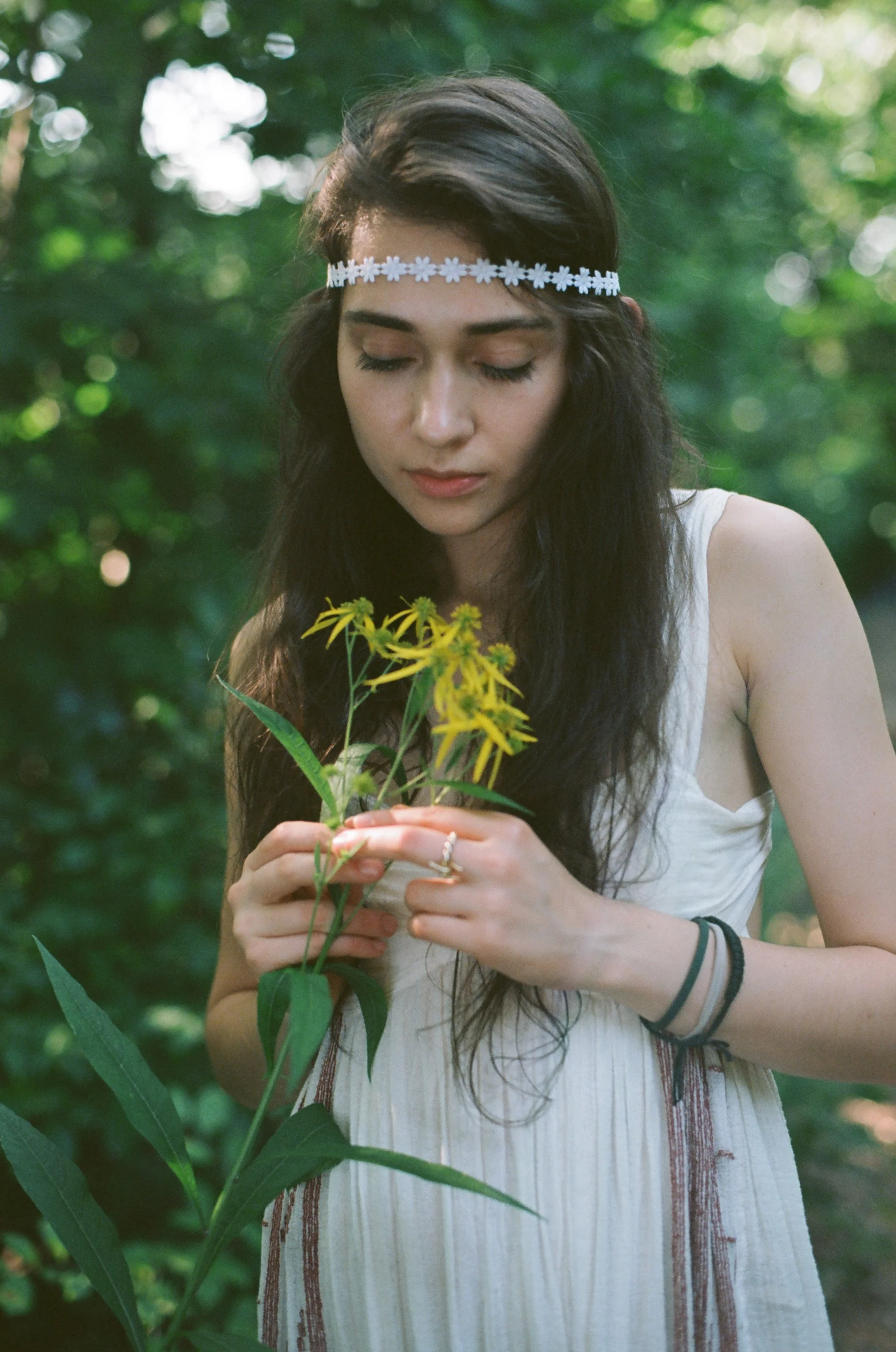 Young woman with long dark hair, wearing a white dress and a daisy floral headband, holding yellow flowers in her hands in a green outdoor setting.