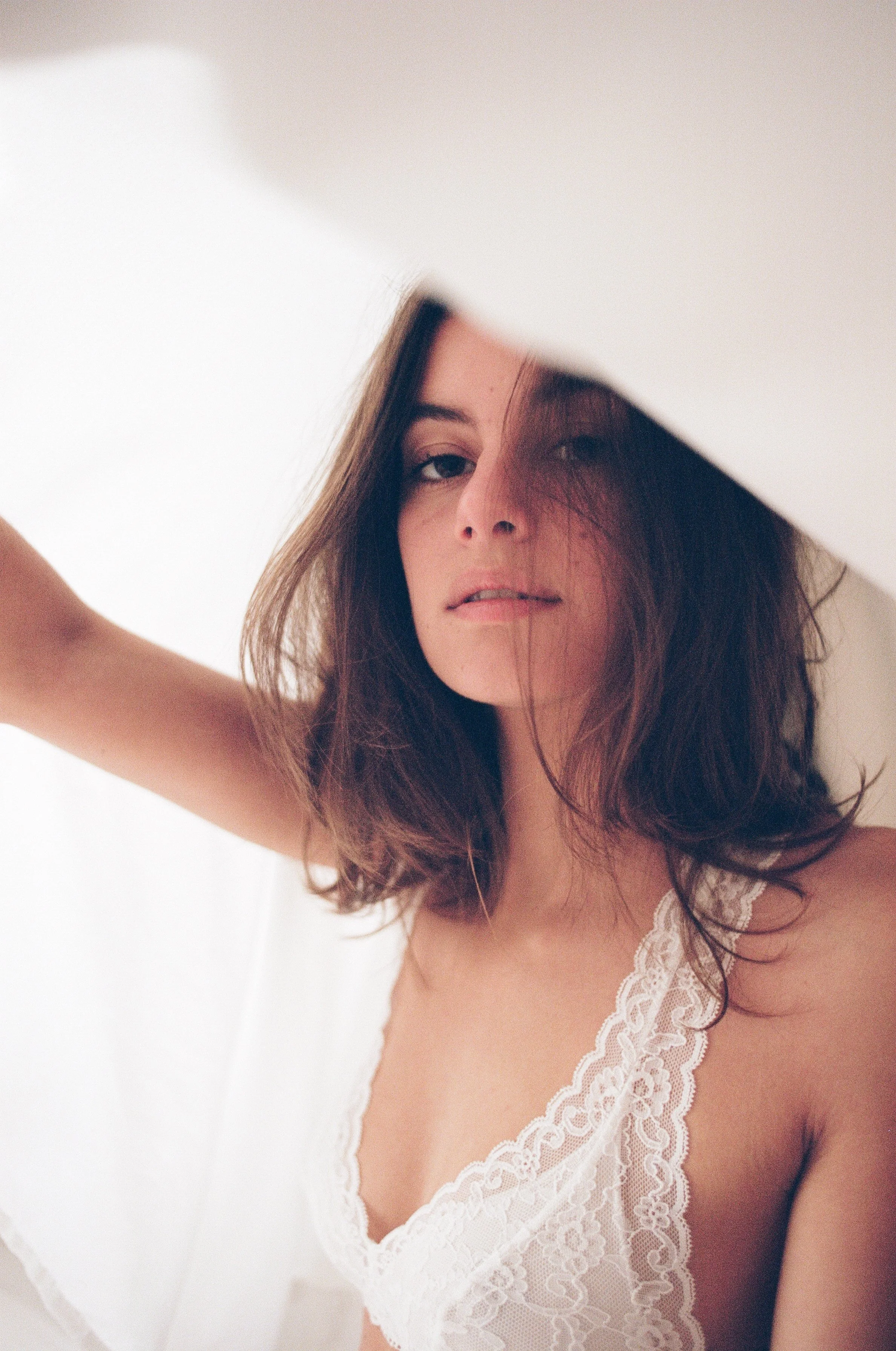 A woman with shoulder-length dark hair and fair skin looking through a gap in white fabric, wearing a white lace top.