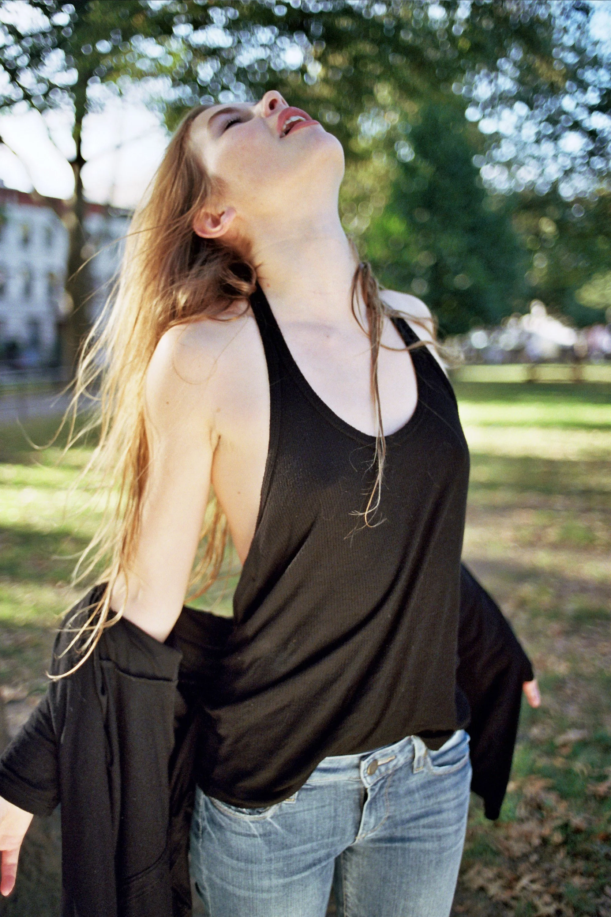 A woman with long, wet hair wearing a black tank top and light blue jeans stands outdoors in a park, tilting her head back with her eyes closed and an expression of enjoyment.