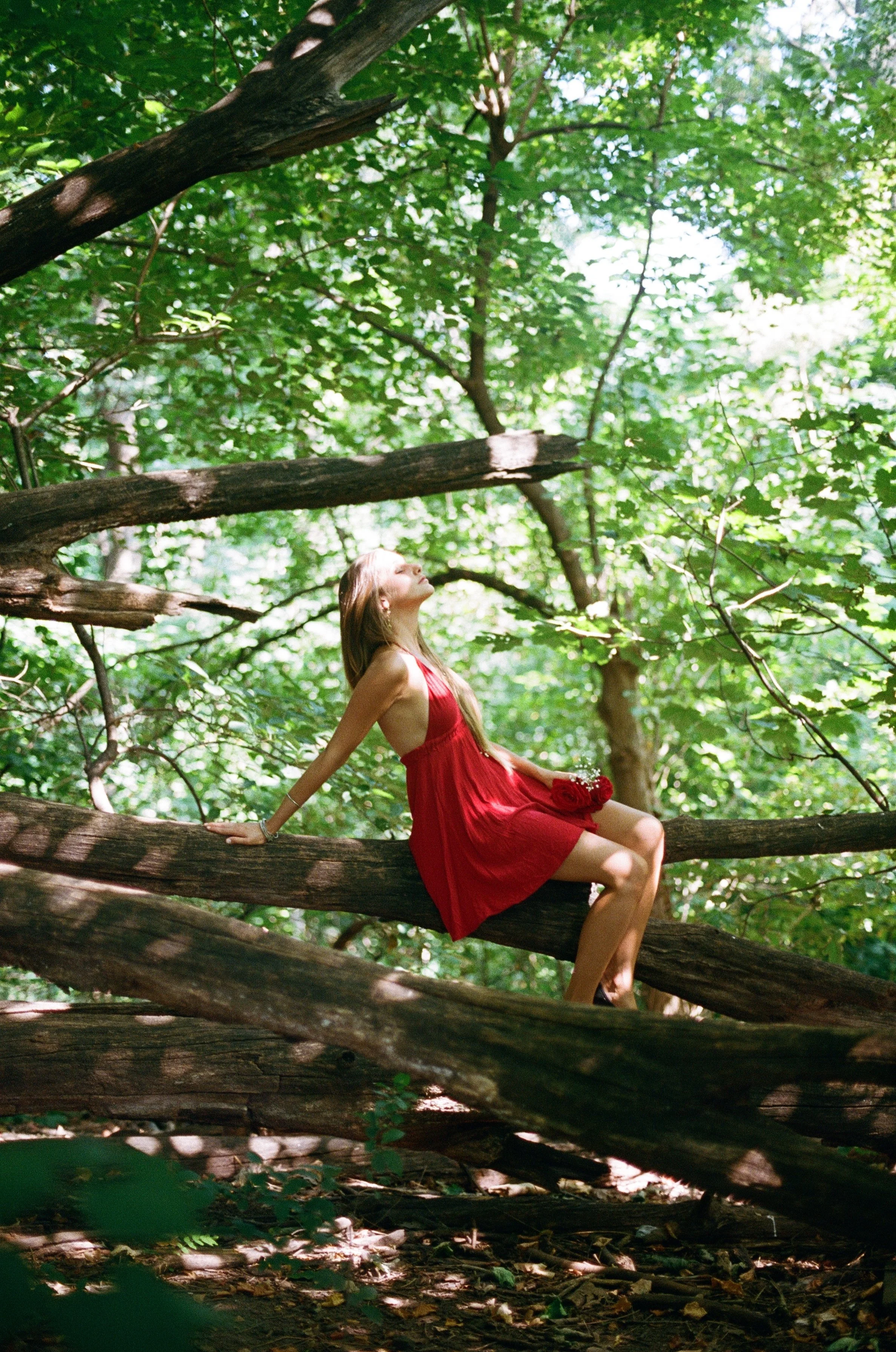 A woman in a red dress sitting on a fallen tree branch in a lush green forest, with her head tilted back and eyes closed.