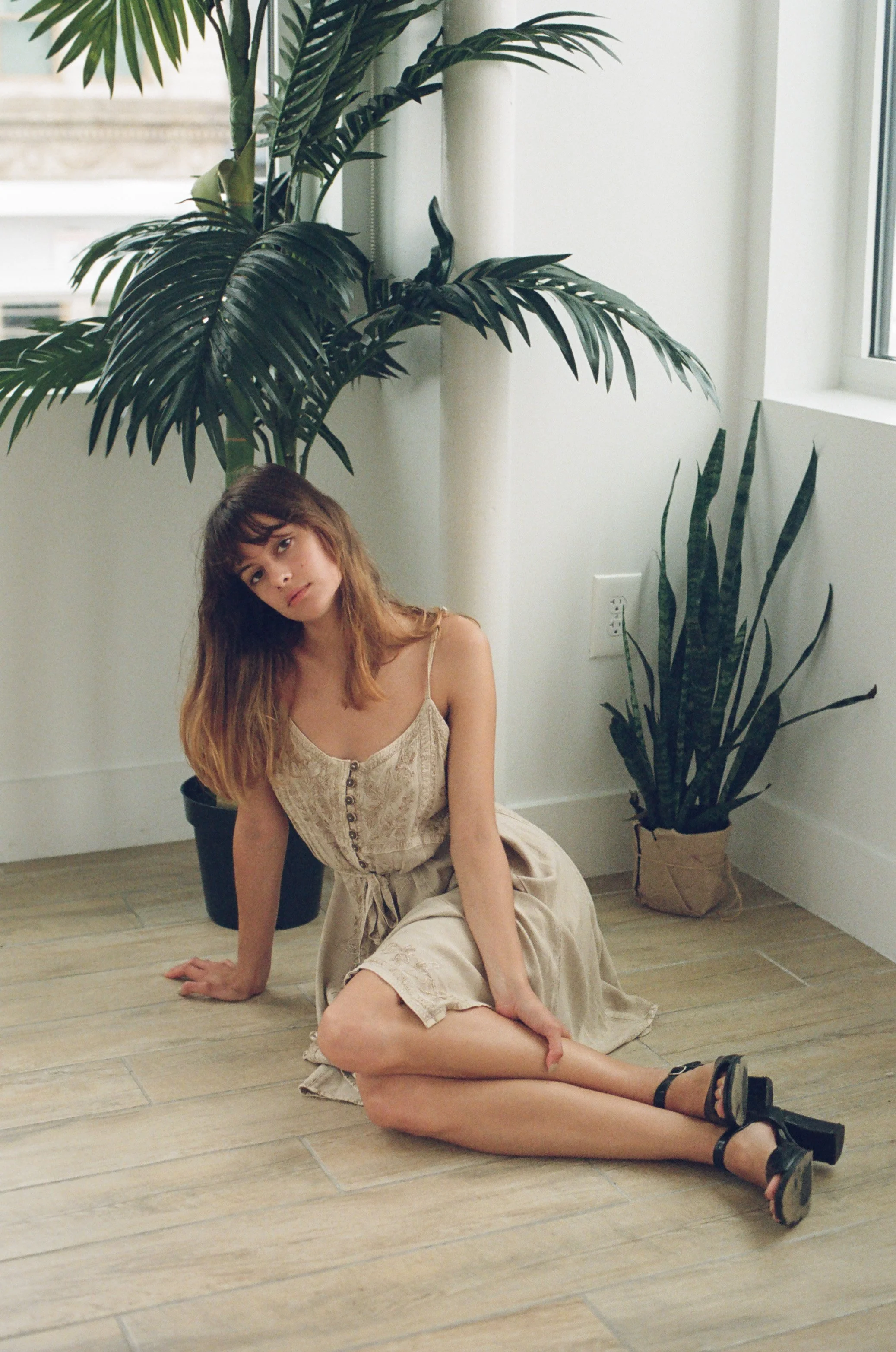 A woman with light brown hair, wearing a beige dress and black high heels, sitting on a wooden floor next to a large potted plant in a bright room with a window.