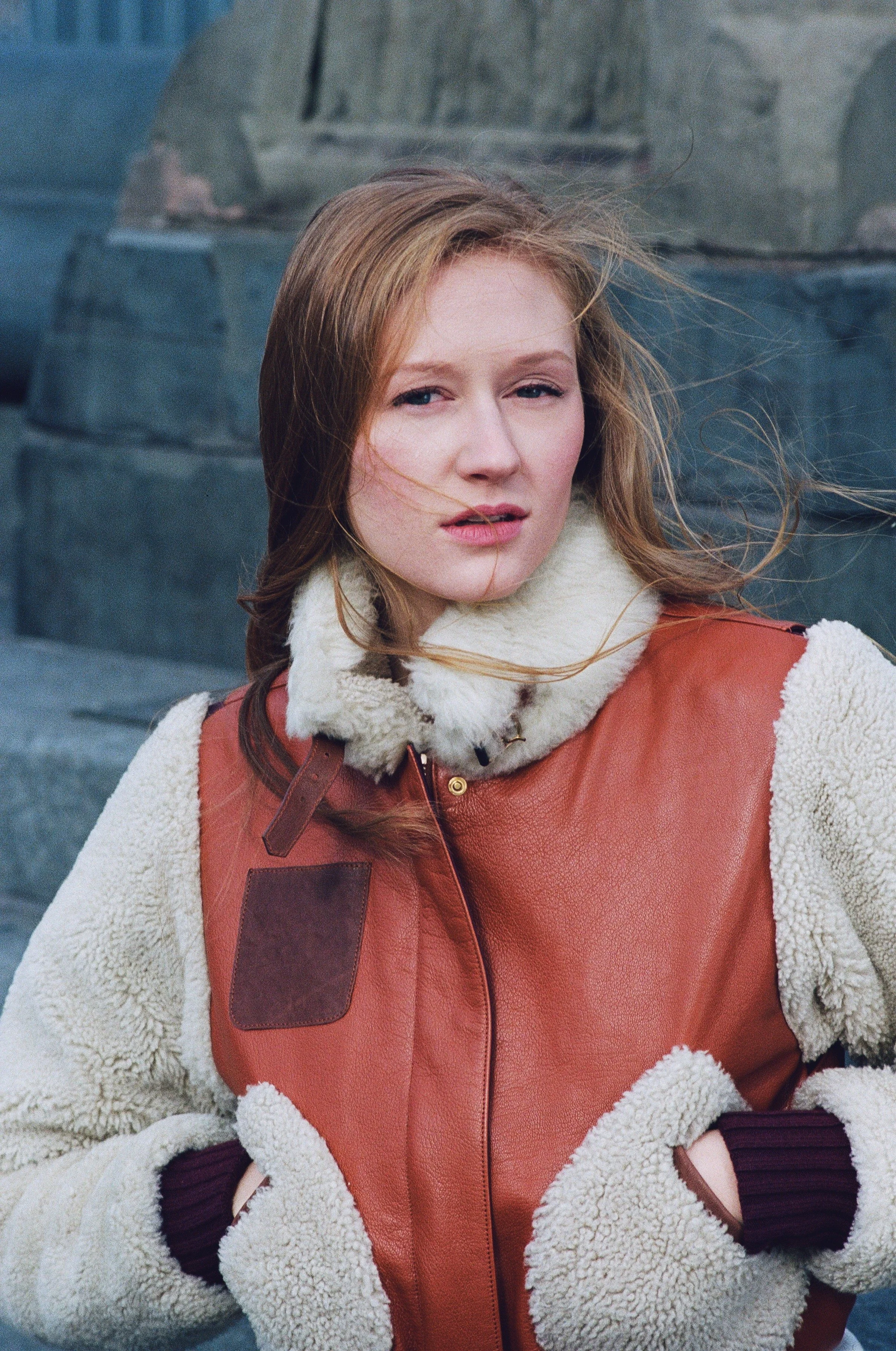  Photo edited by Mary Perrino.A young woman with reddish hair and fair skin standing outdoors with windblown hair, wearing a warm cream-colored shearling-lined jacket with a rust-colored leather front.