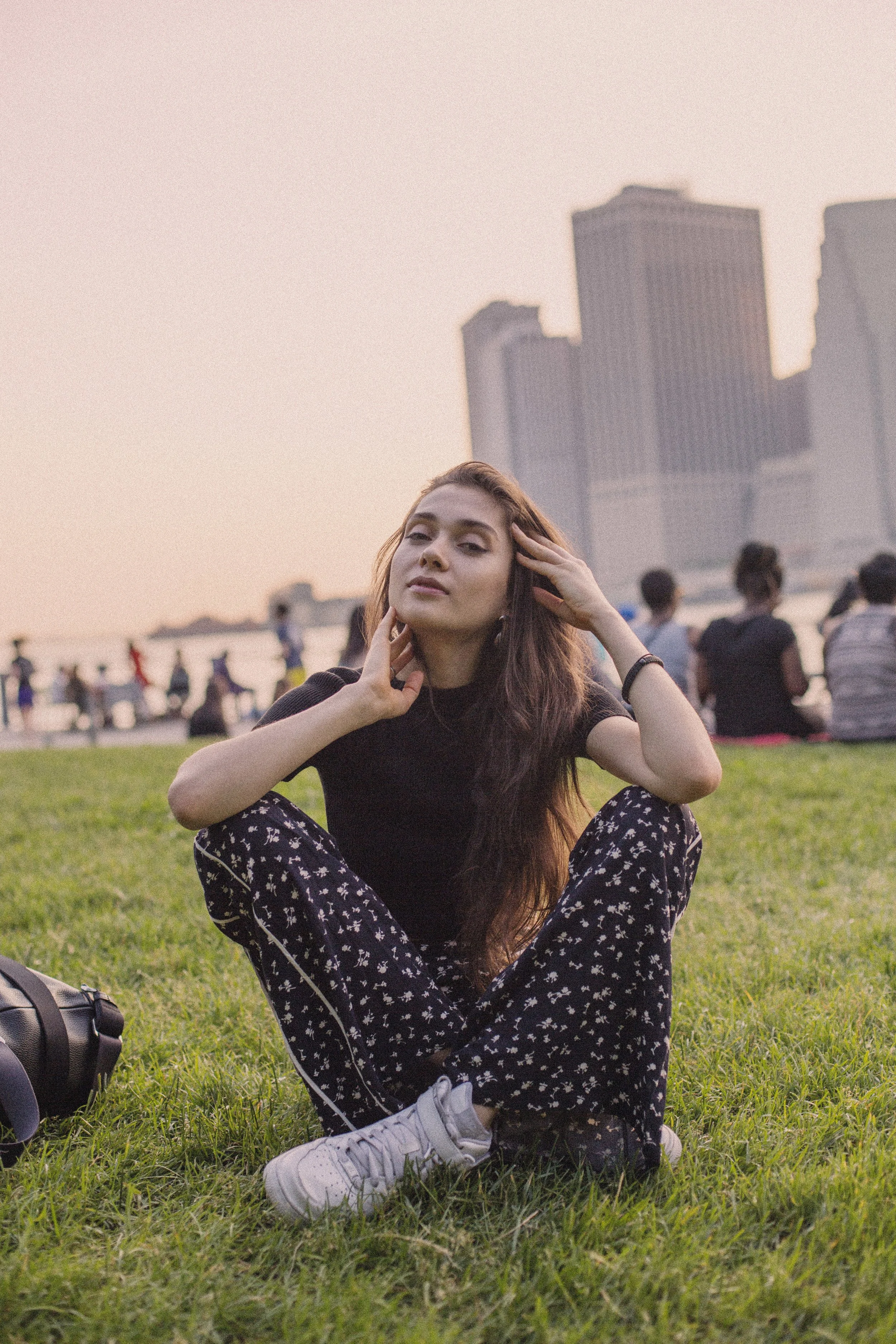 A young woman sitting cross-legged on grass at sunset, with a city skyline and people in the background.