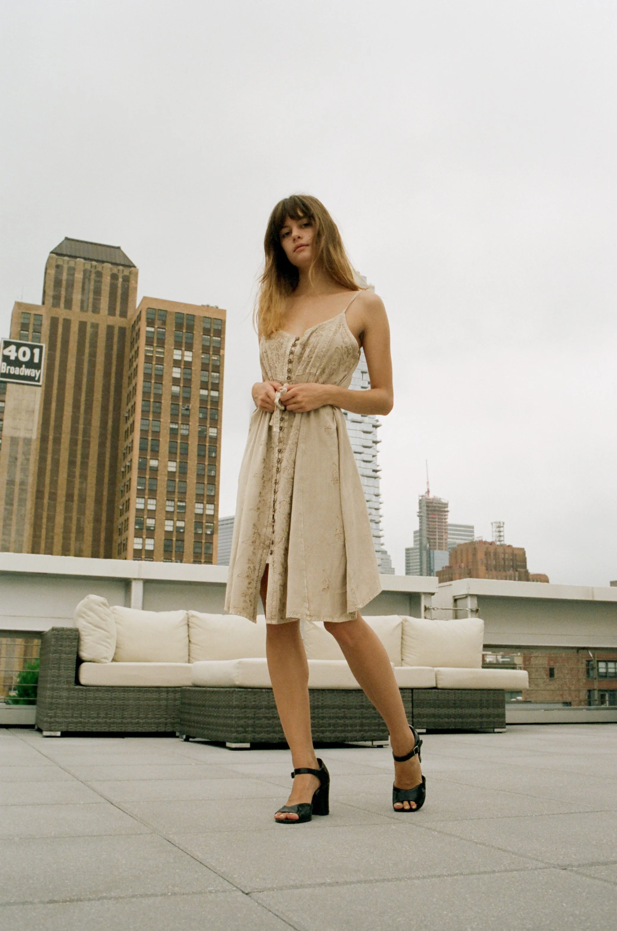 Young woman in a sleeveless beige dress and high heels standing on a rooftop with city skyscrapers in the background.