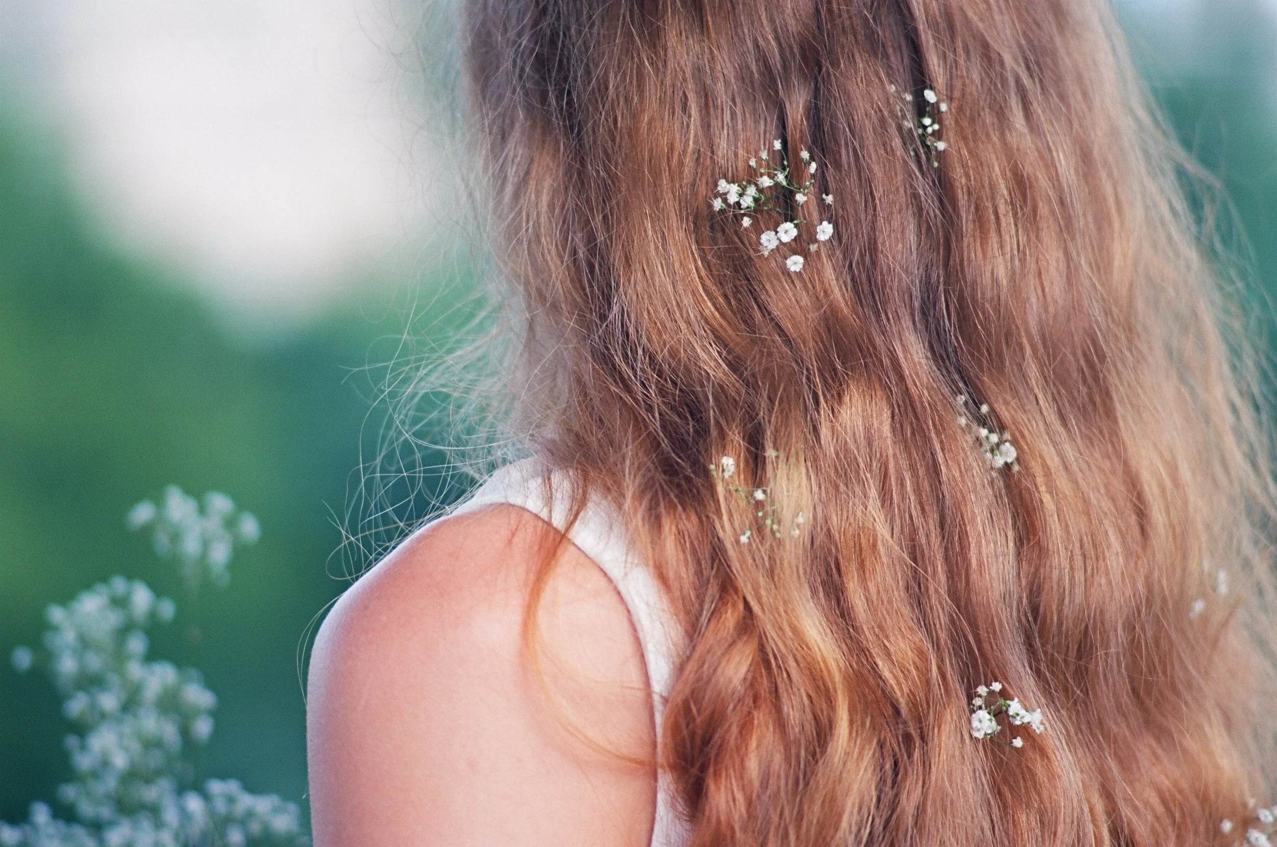 Close-up of a woman with long, wavy red hair decorated with small white flowers, wearing a sleeveless white top, outdoors on a sunny day.