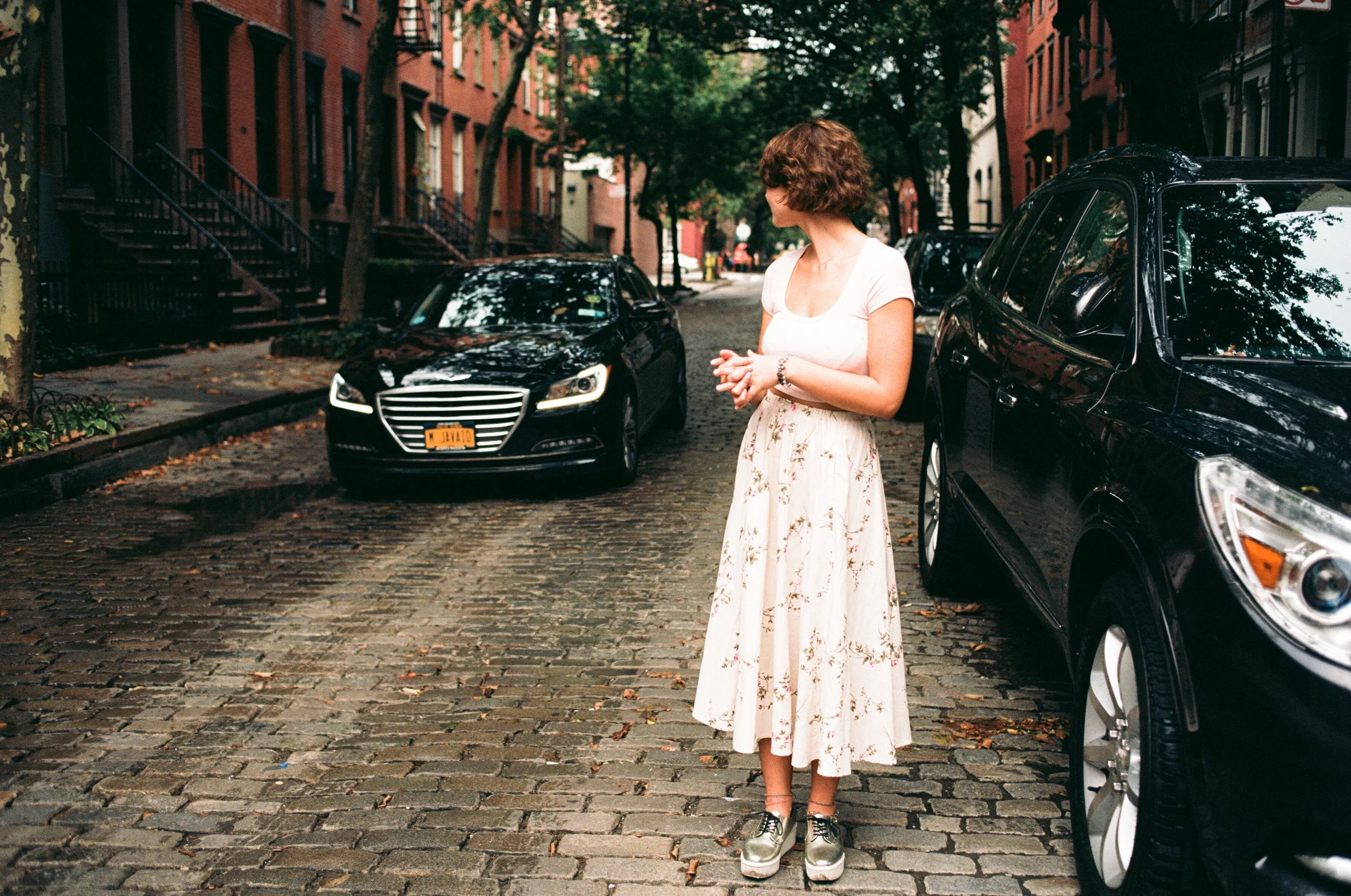 Woman in a white top and floral skirt standing on a cobblestone street with parked black cars and colorful townhouses.