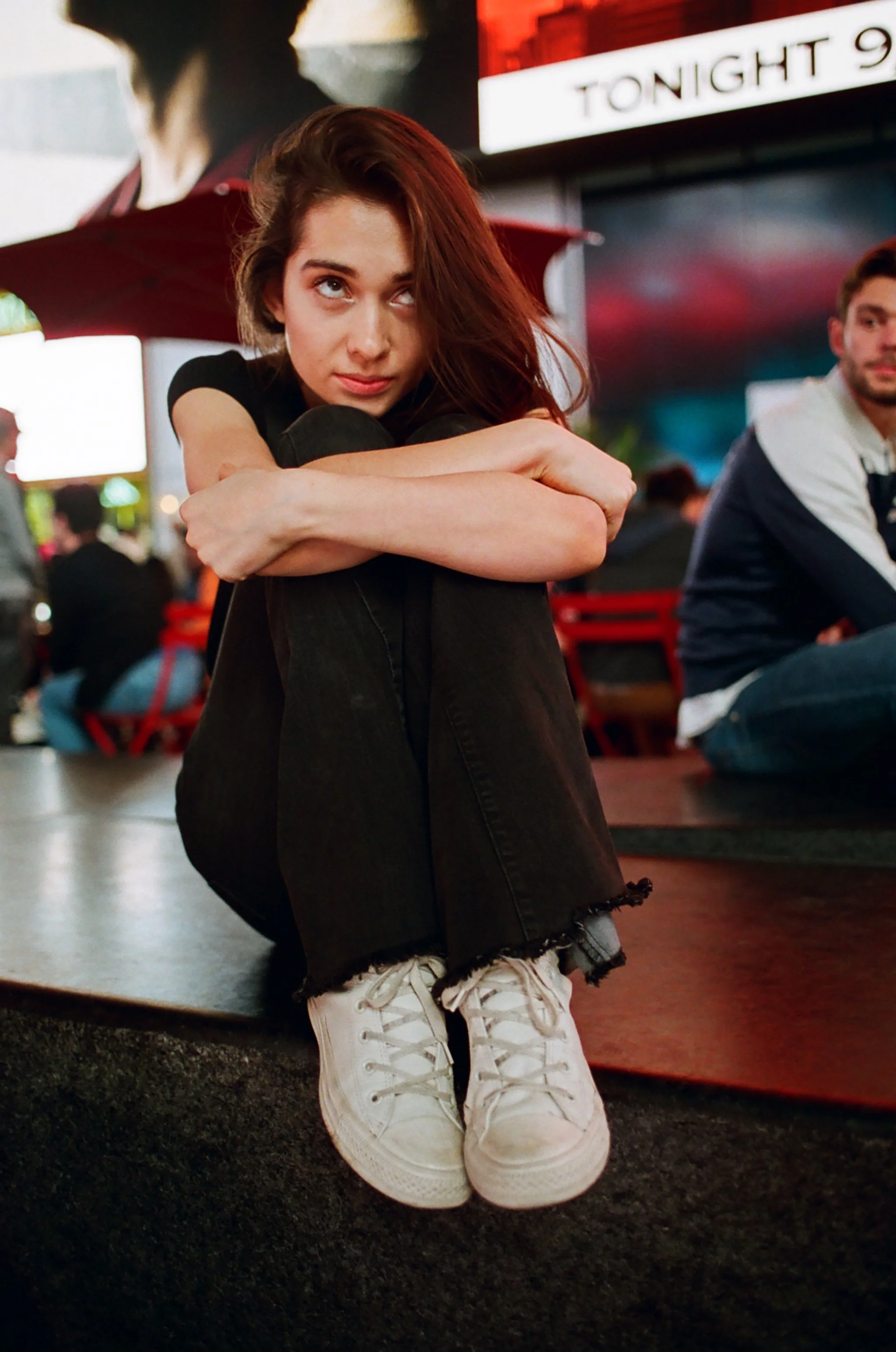 A young woman with brown hair sitting on a curb in an urban setting, hugging her knees, with a serious expression. In the background, there are people sitting and a lit screen displaying 'TONIGHT 9'.