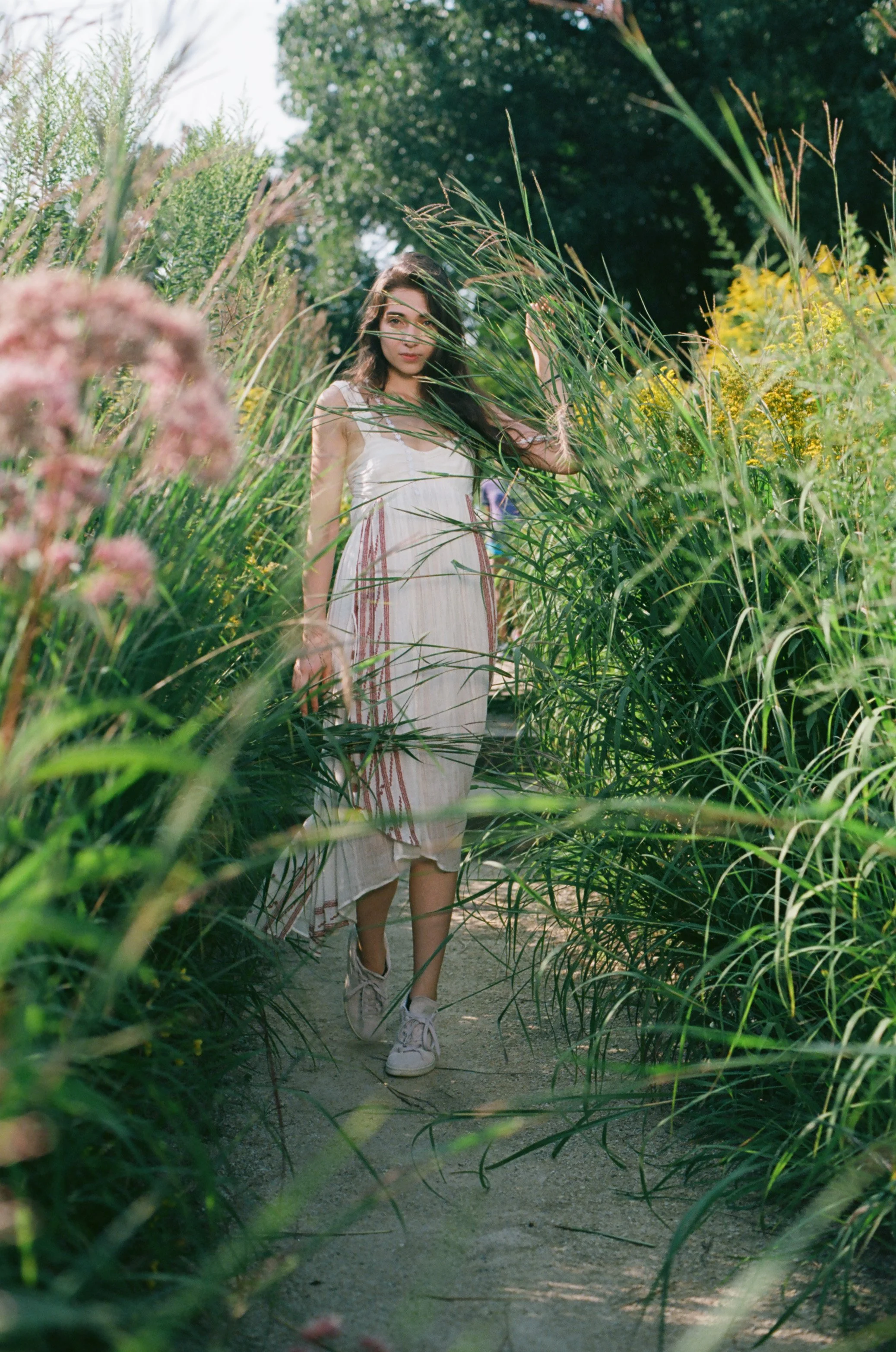 A young woman with long dark hair wearing a white dress and sneakers walking through a lush green garden pathway surrounded by tall plants and flowers.