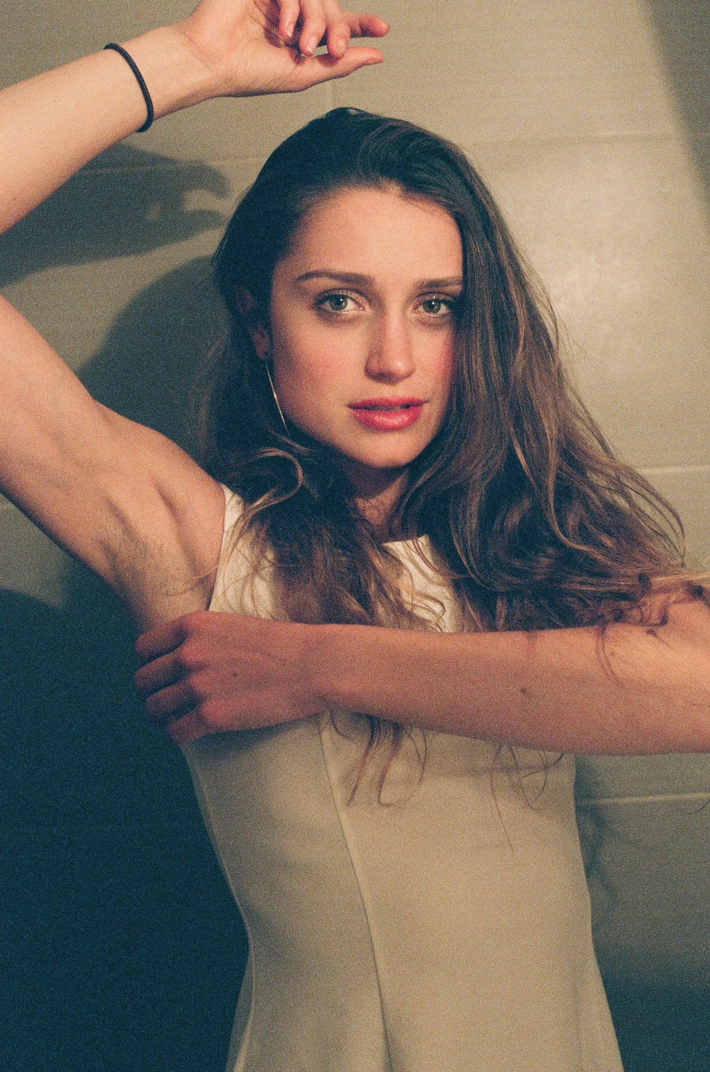 A woman with long, wavy brown hair and fair skin is leaning against a wall, looking directly at the camera with a slight smile. She has one arm raised above her head and the other resting on her shoulder, wearing a sleeveless top and a black hair tie