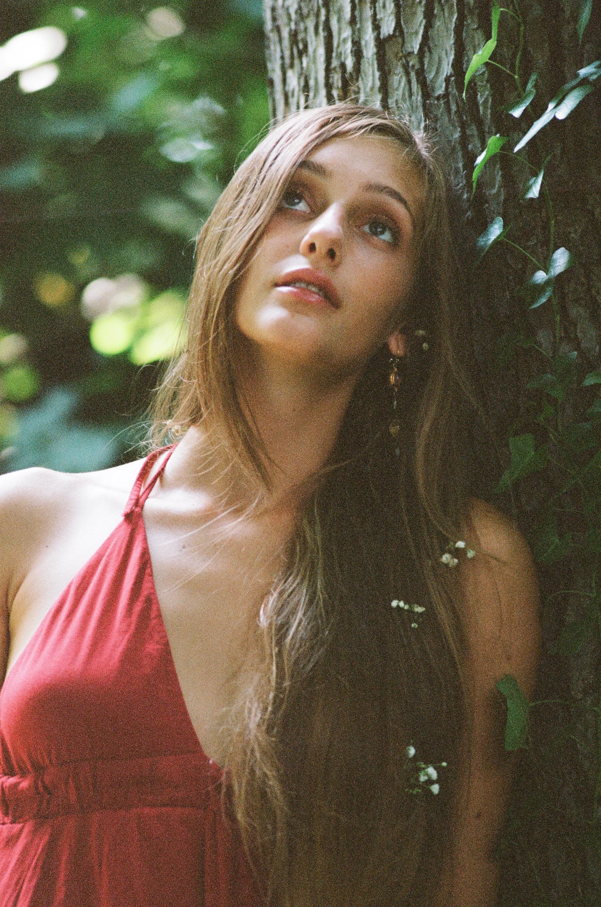 Young woman with long brown hair and blue eyes wearing a red dress, leaning against a tree in a green forest, looking upward.