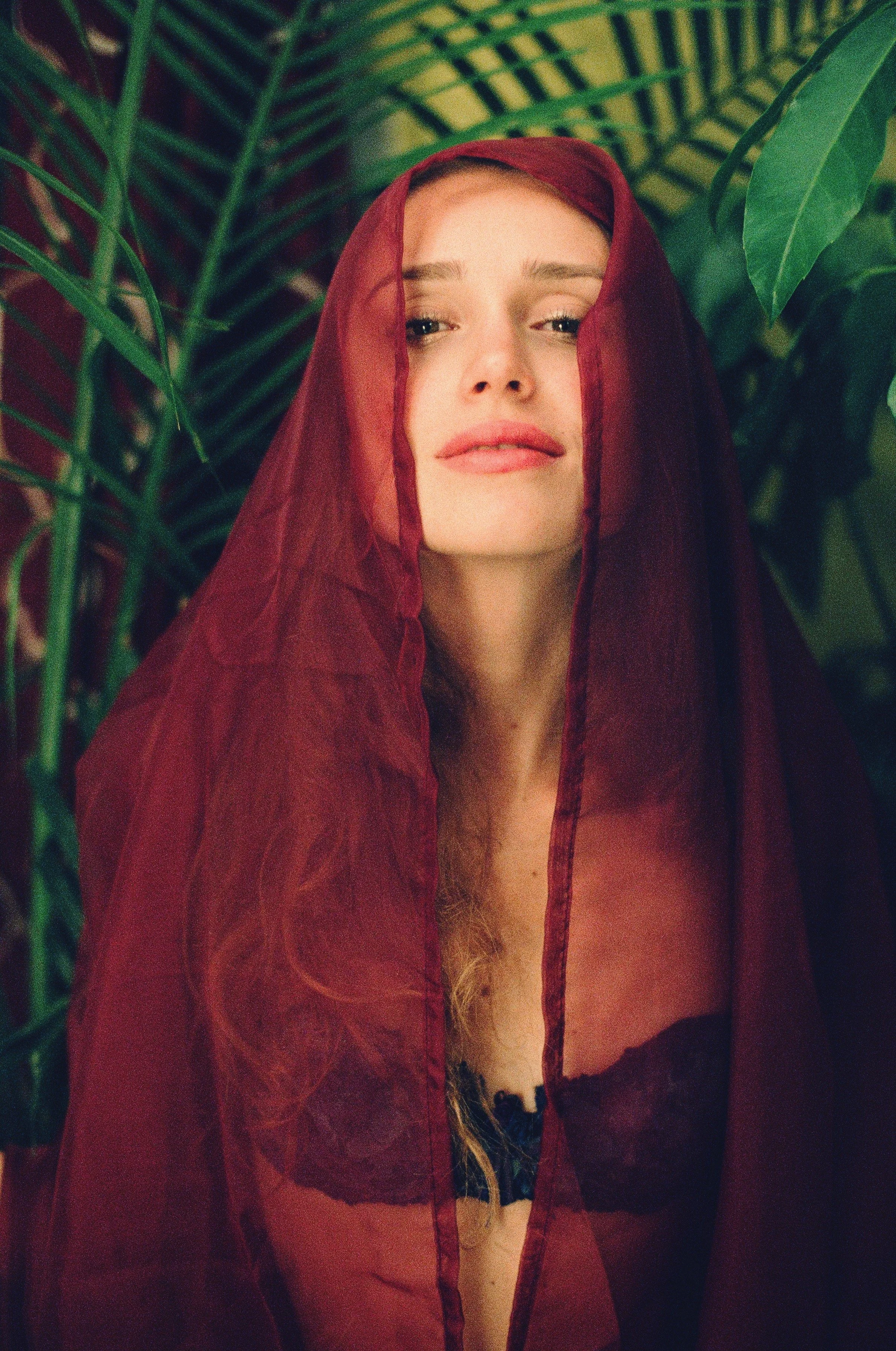 A woman with fair skin and blonde hair partially visible, wearing a sheer black lace bra and a red veil, standing among green tropical leaves. Photo edited by Mary Perrino.