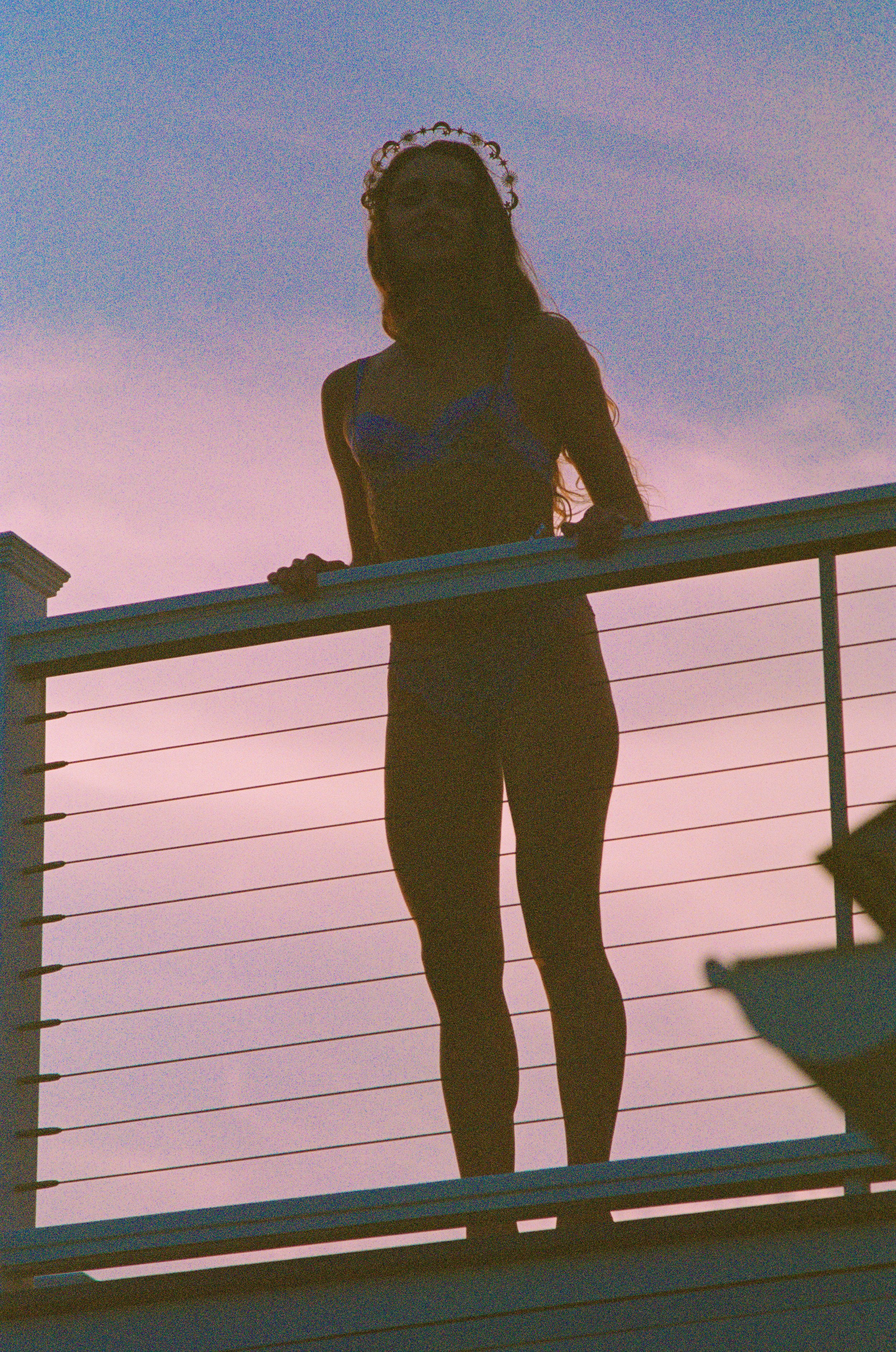 Silhouette of a woman wearing lingerie and a tiara, standing on a balcony against a twilight sky. Photo edited by Mary Perrino.