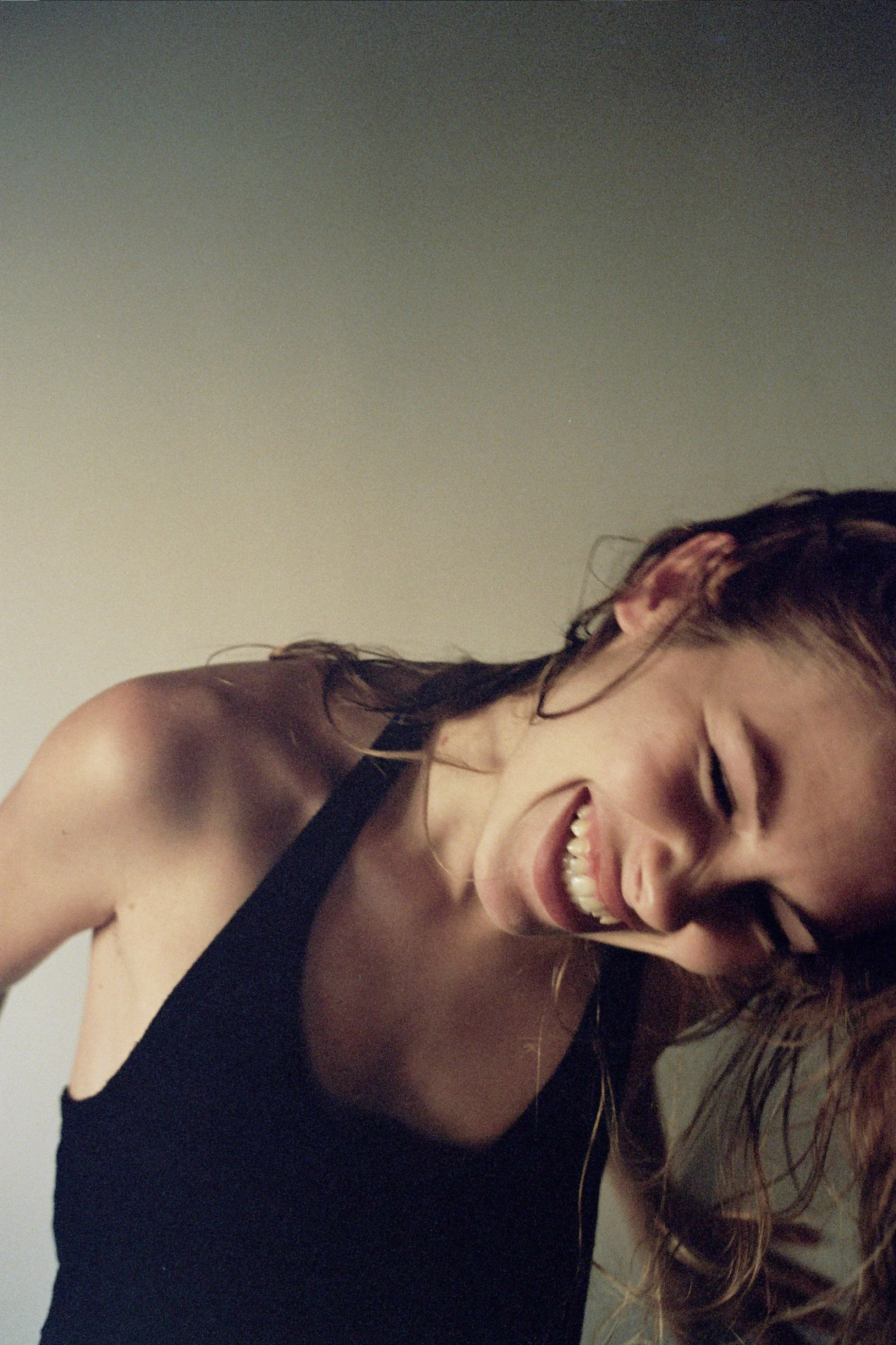 A woman smiling with her head tilted to the side, wearing a black tank top, and with light brown hair, against a plain background.