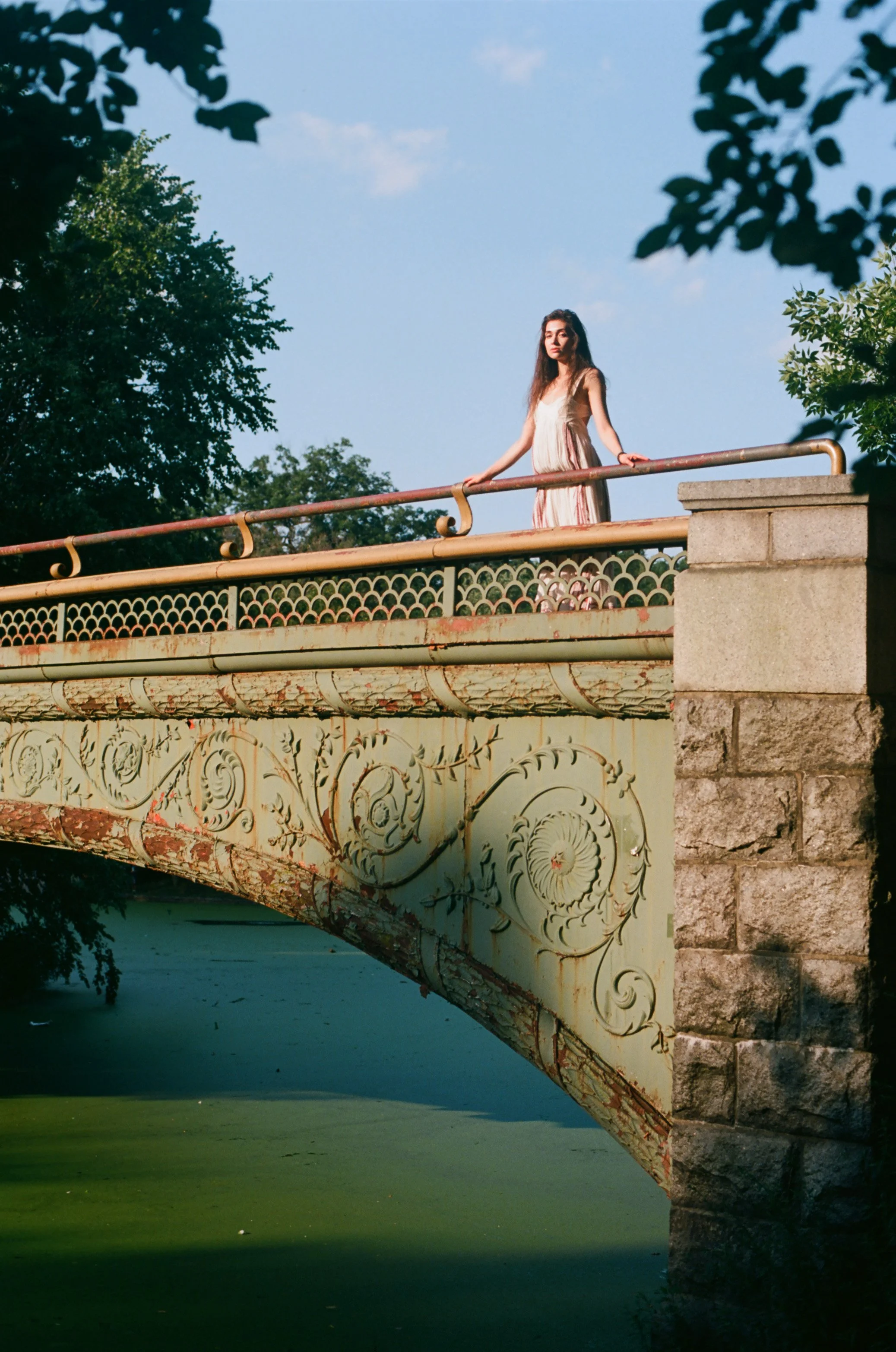 Young woman standing on an ornate stone bridge with iron railings, with trees and a blue sky in the background. Photo edited by Mary Perrino.