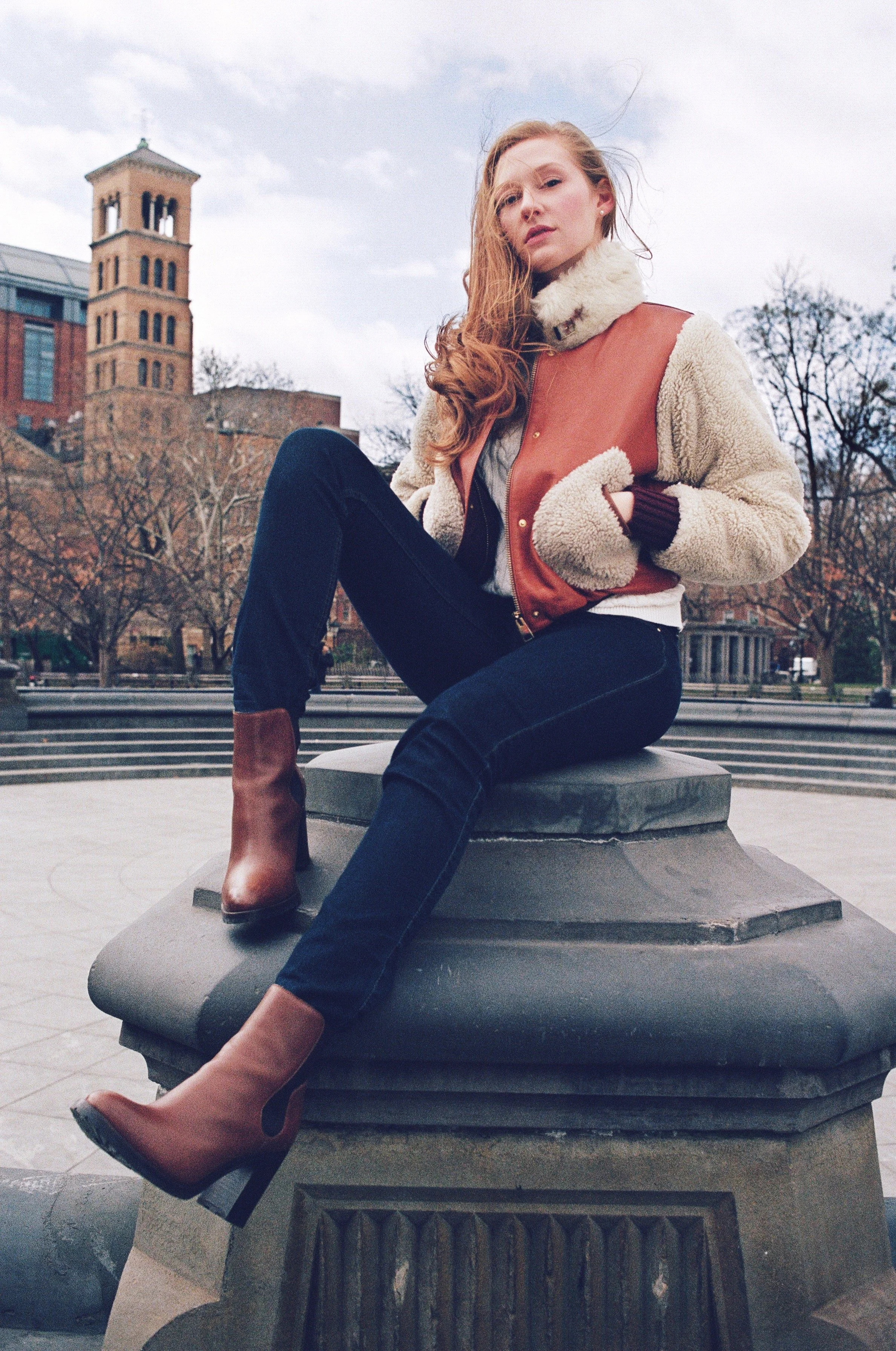 A young woman with long red hair sitting on a stone structure outdoors with an urban background of trees, buildings, and a tower, wearing a sherpa-lined jacket, black jeans, and brown ankle boots.